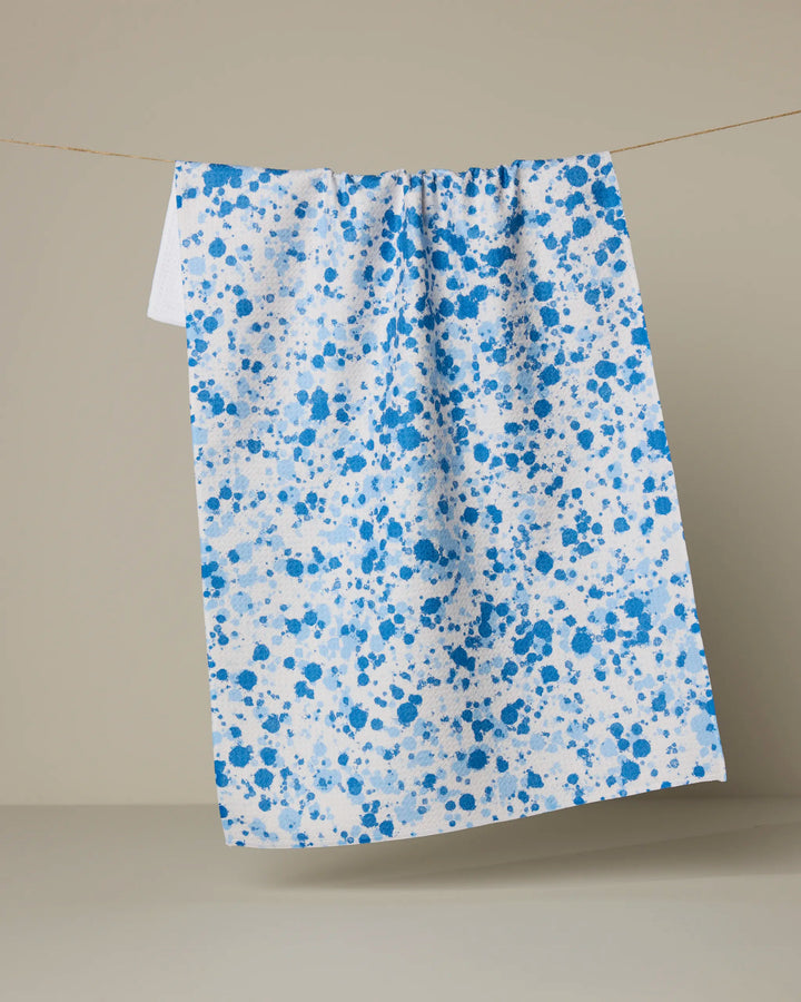 The Indigo Splash tea towel by Geometry, crafted from recycled materials with a blue paint splatter design, hangs on a clothesline against a plain beige background.