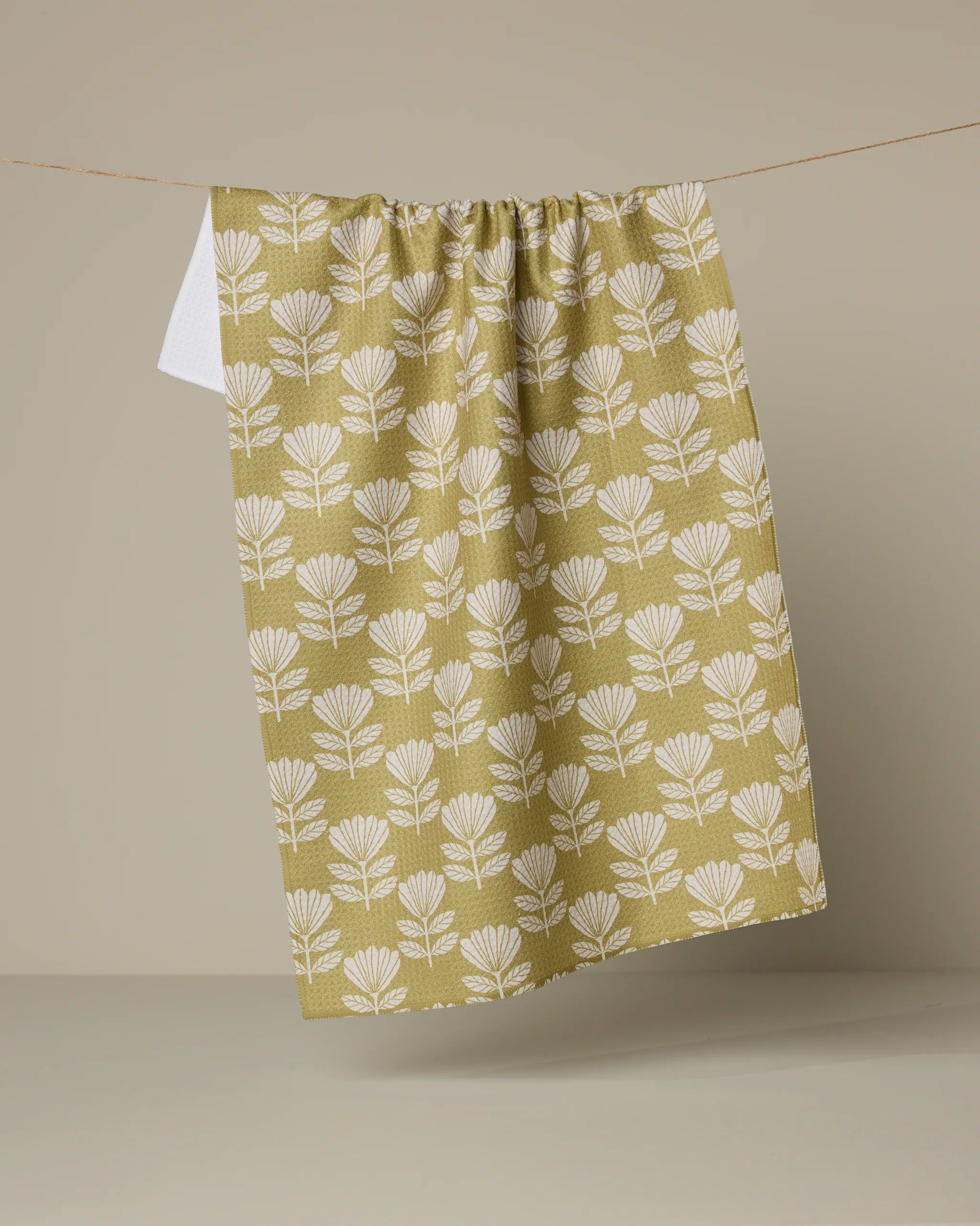 The Anna Christian Ingrid kitchen towel, made from recycled materials and featuring a white floral pattern on green, hangs on a line against a beige background.