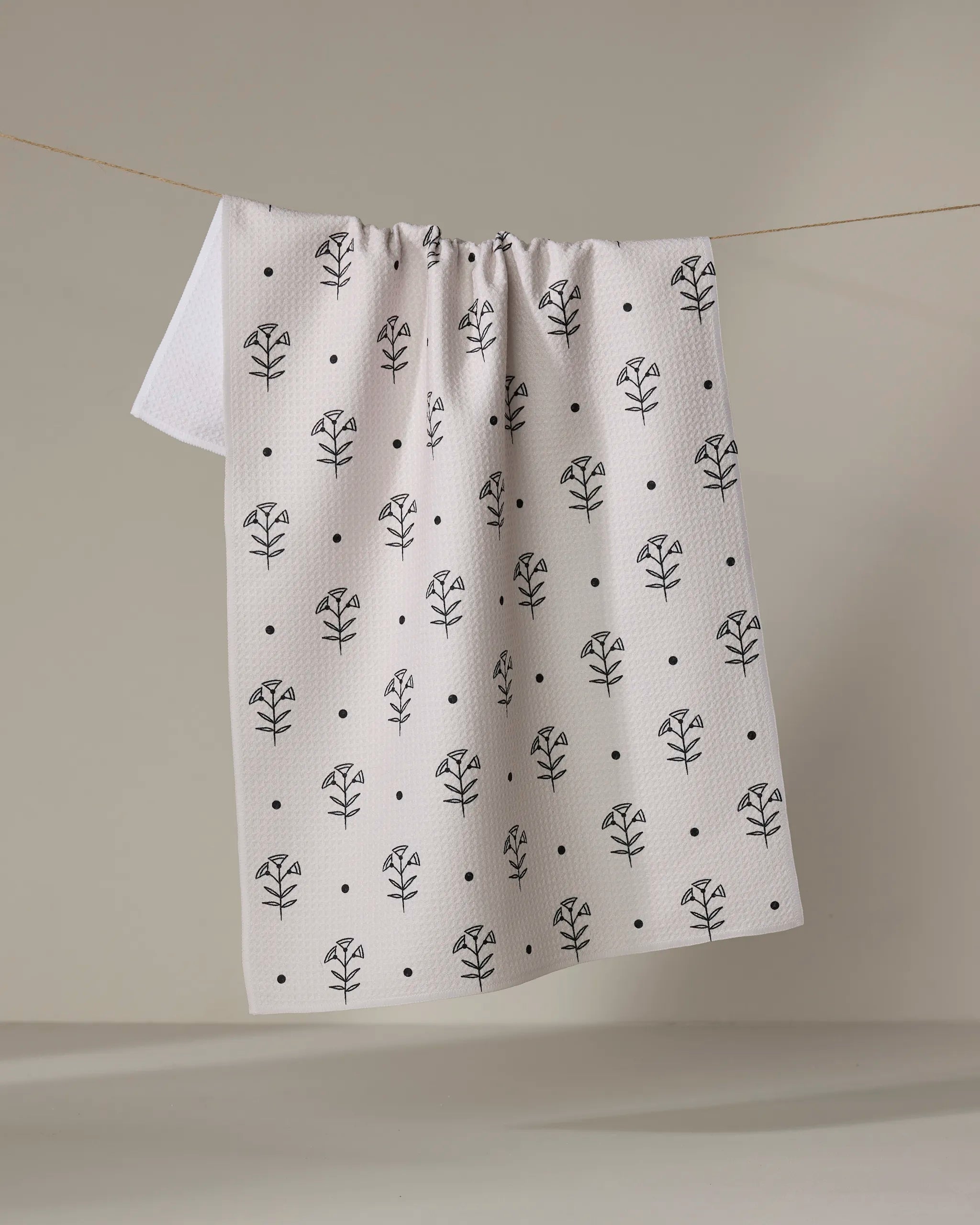 The Cole Strem "Inked Bloom" dish towel, featuring a black floral and dot pattern on white, is shown hanging over a clothesline against a neutral background.