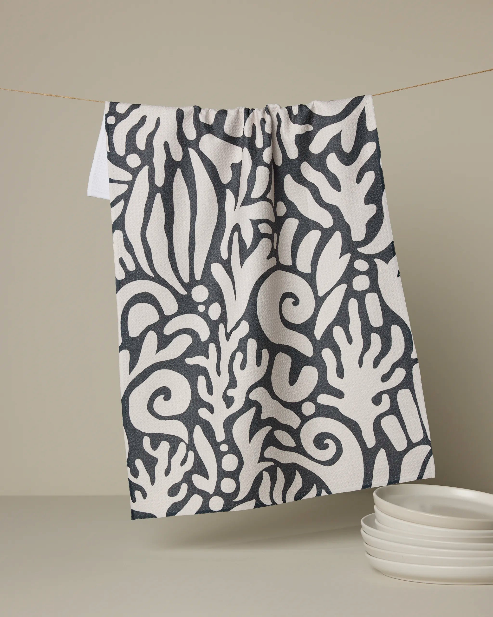 The Geometry Inkwell Reef tea towel, featuring a bold black and white abstract botanical pattern, hangs on a line above neatly stacked white plates against a neutral background.