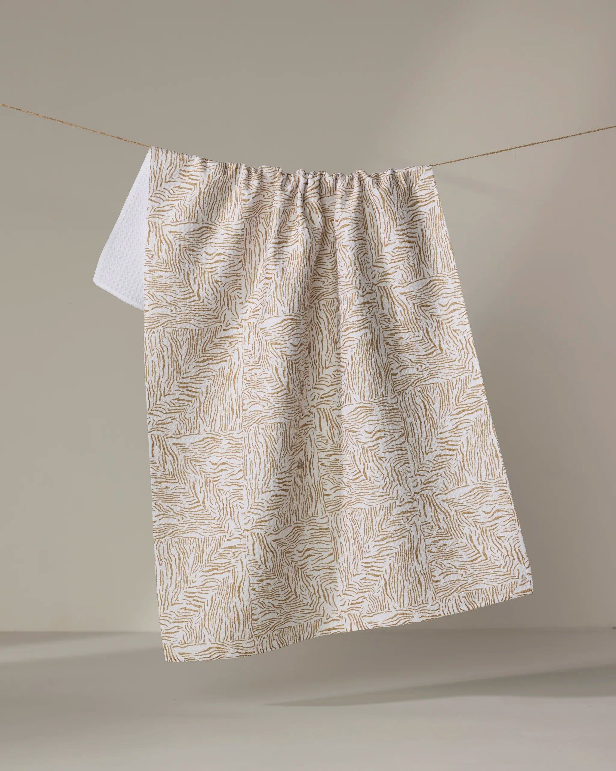 The "Inlay" recycled kitchen towel by Studio Jeannot, featuring a beige and white abstract wavy pattern, hangs on a clothesline against a neutral background.