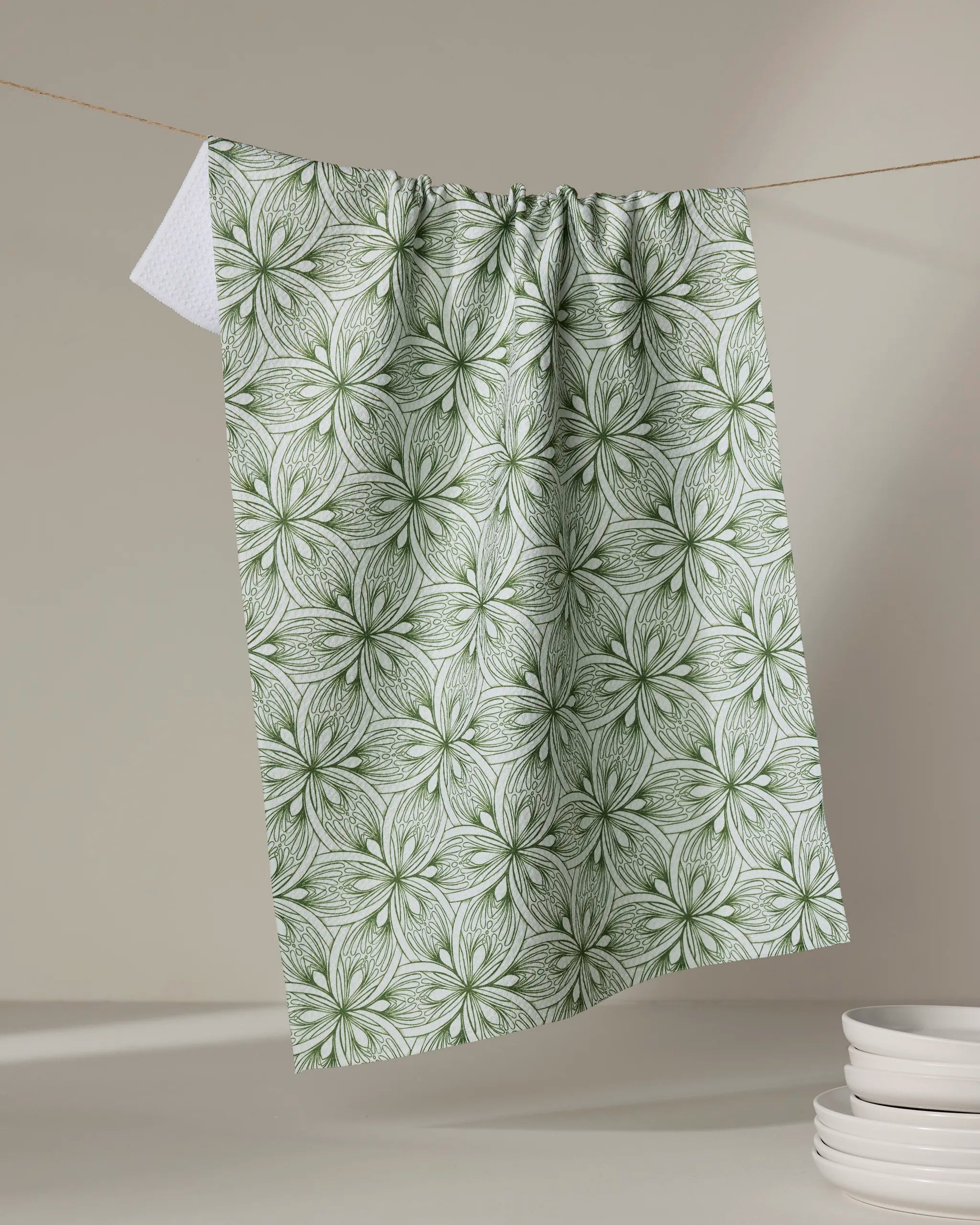 The Marta Harvey "Interlaced Petals" tea towel, featuring a green and white repeating floral pattern, hangs on a string against a neutral background with white plates stacked in the lower right corner.