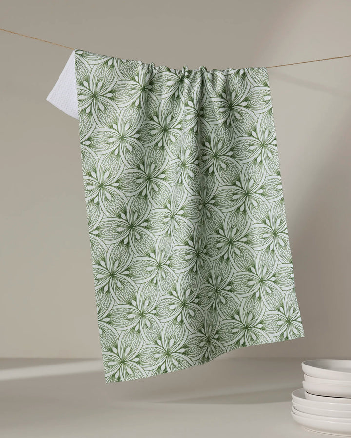 The Marta Harvey "Interlaced Petals" tea towel, featuring a green and white repeating floral pattern, hangs on a string against a neutral background with white plates stacked in the lower right corner.