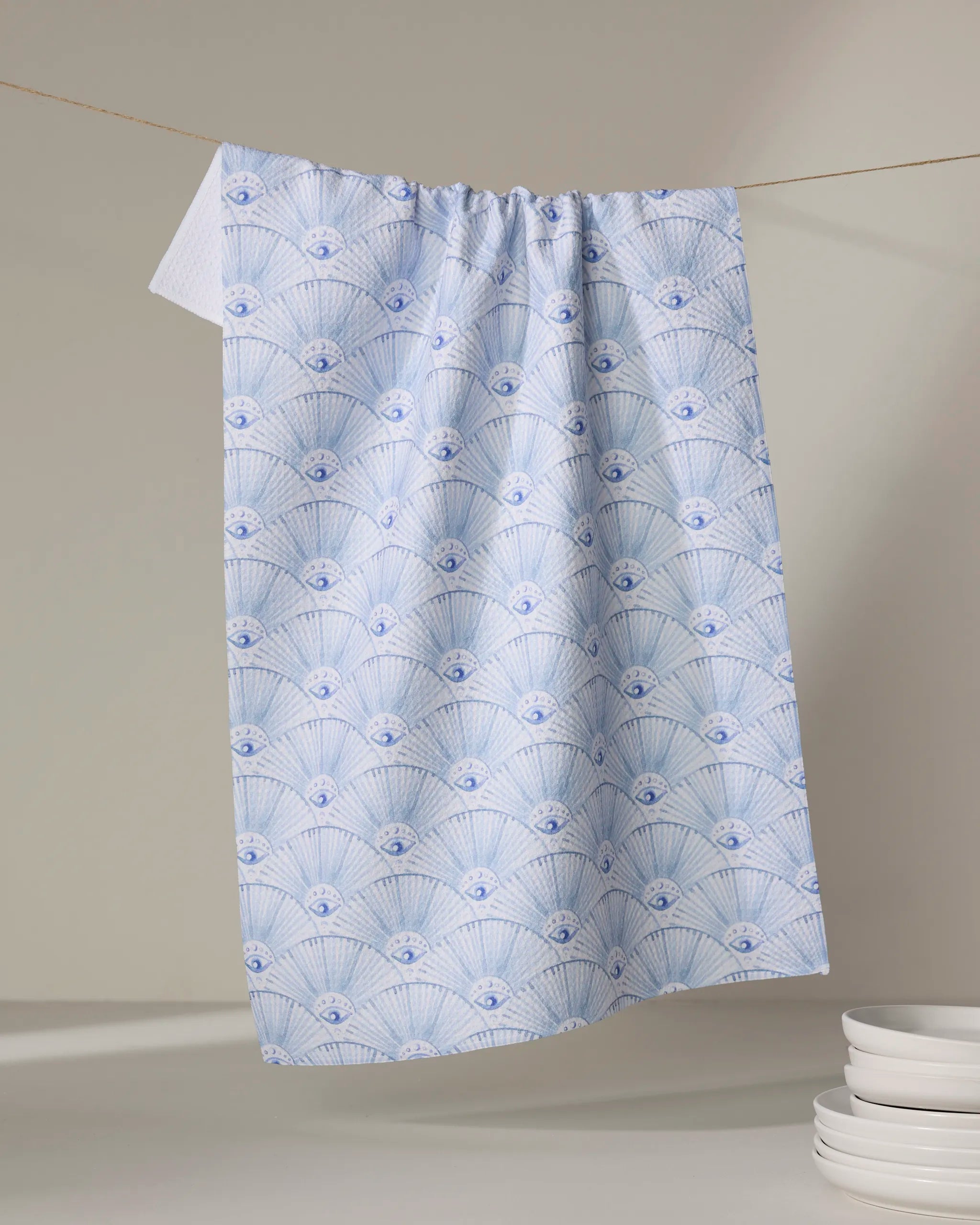 The "Into The Mystic" tea towel by Maayan Alper-Swan, crafted from recycled materials, displays a light blue and white scallop shell pattern and hangs on a line with stacked white dishware shown in the bottom right.