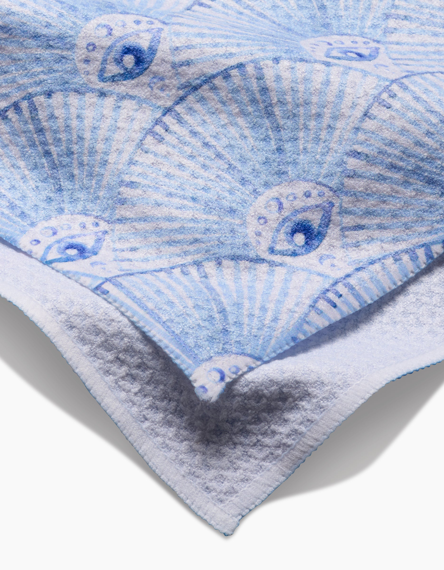 A close-up of the soft, white Maayan Alper-Swan Into The Mystic tea towel, featuring a blue seashell and eye pattern on Geoweave fabric made from post-consumer recycled materials, highlighting its detailed texture and folded corners.