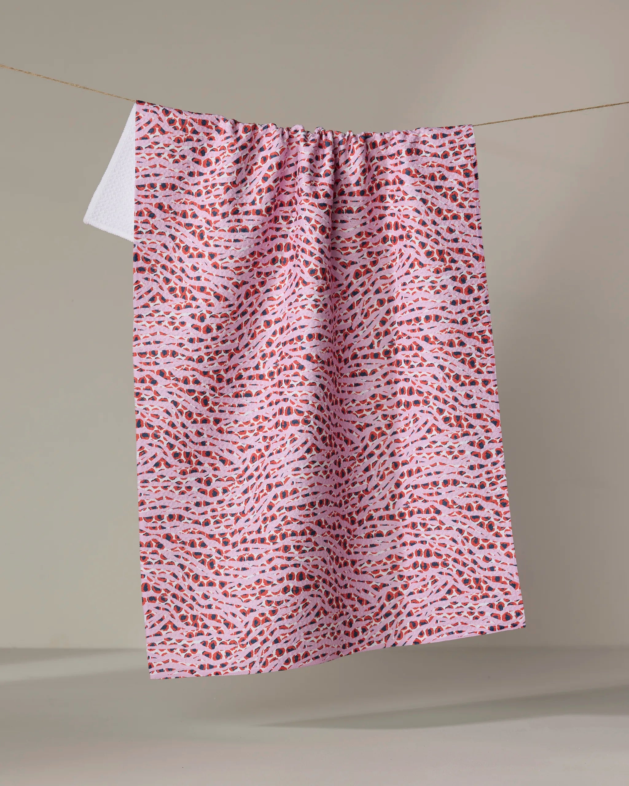 The Happy Hemera "Into the Wild" towel, in pink and white with small red and purple abstract shapes, hangs neatly on a clothesline against a plain, light background.