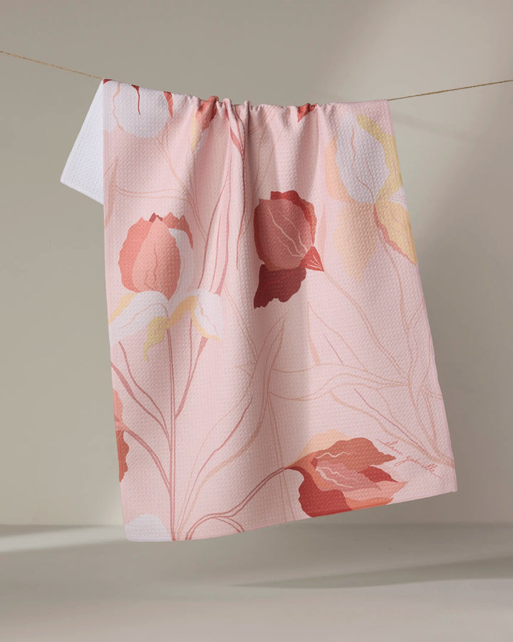 The Iris dish towel by Elana Gabrielle, featuring a large floral pattern in red, pink, yellow, and white on light pink, hangs on a string against a neutral background.