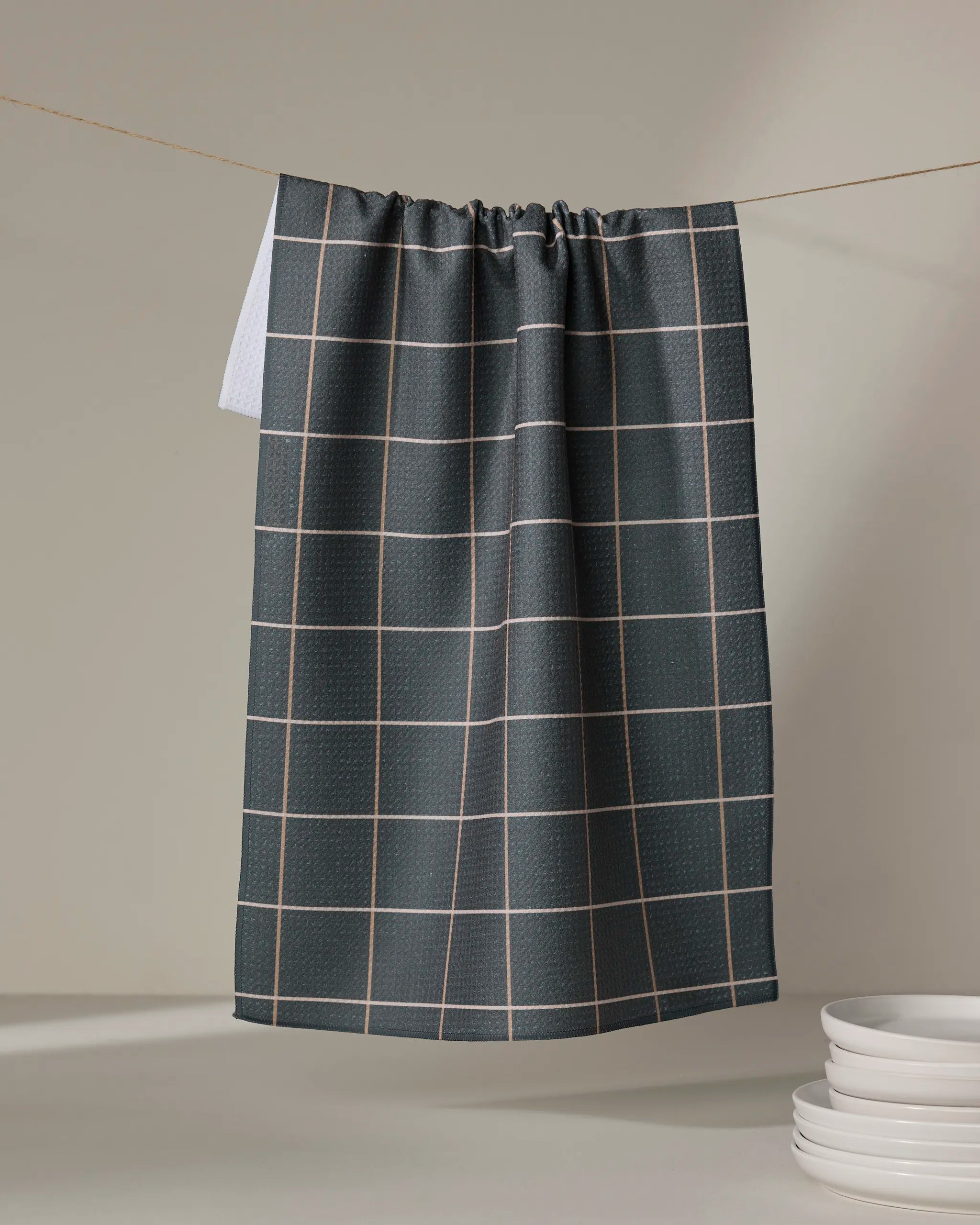 The Kelsi Fullmer Iron Grid kitchen towel, in dark gray with a white grid pattern, hangs on a line with a stack of white plates beside it against a neutral background.