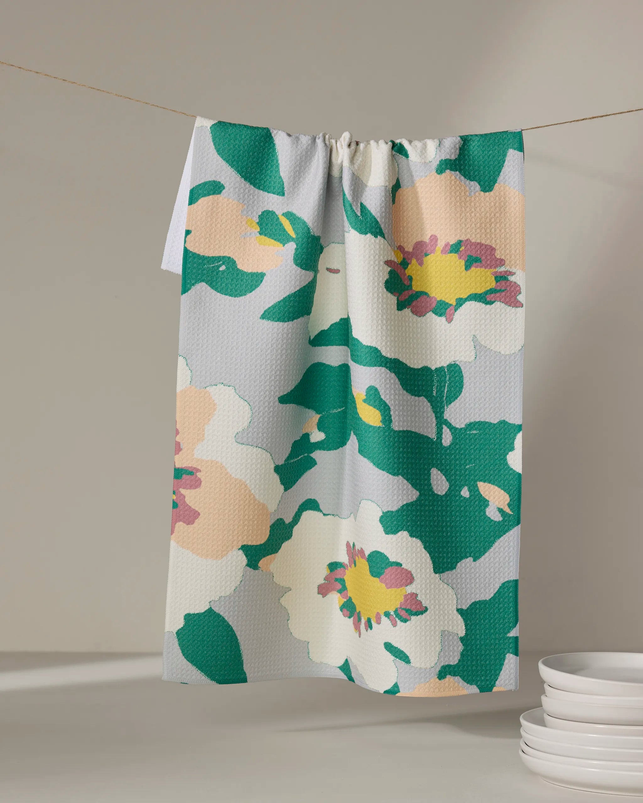 The Geometry Isla tea towel with a quick-dry fabric and large floral print in green, yellow, pink, and white hangs on a string; white plates are stacked nearby.
