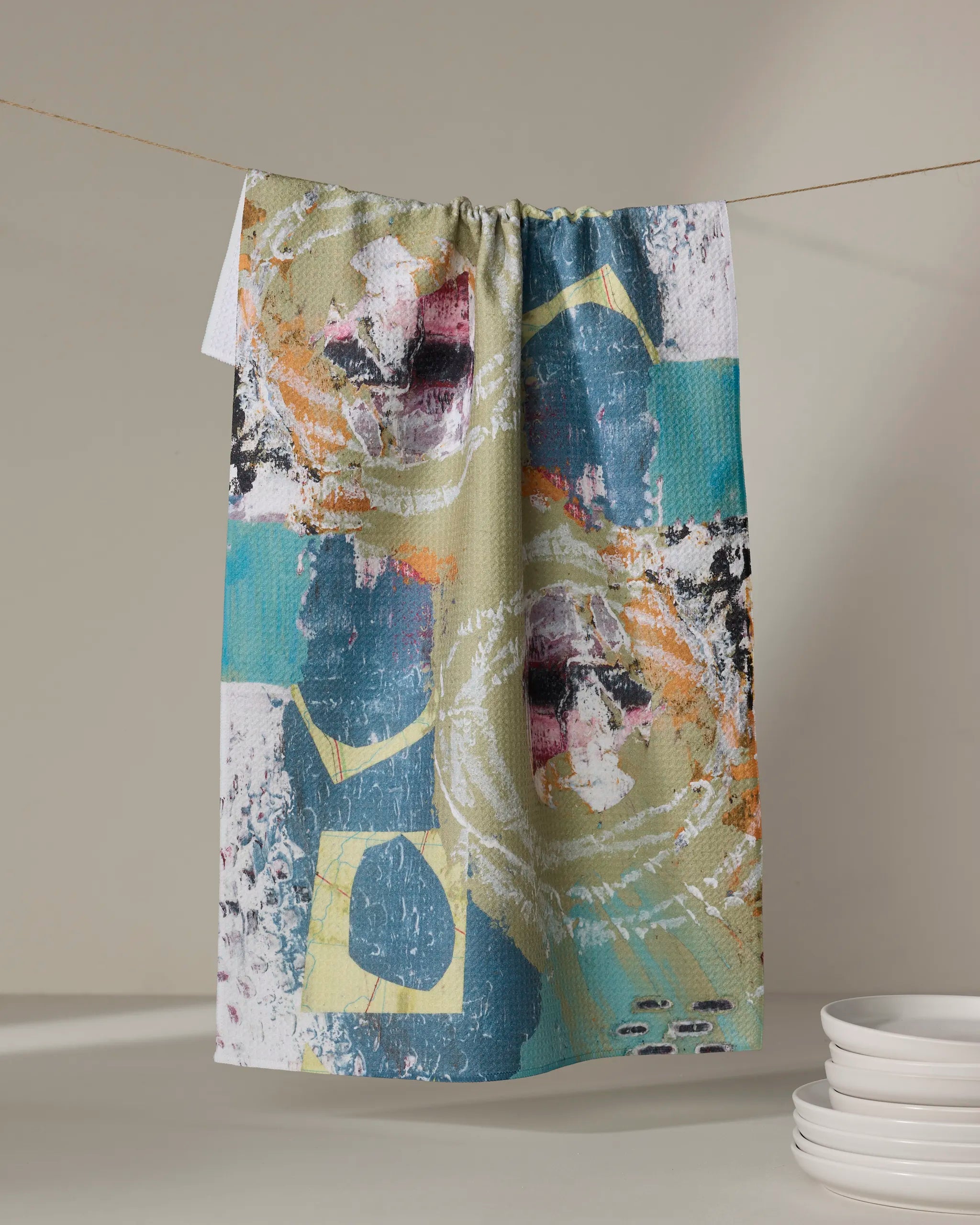 Katherine Berkeley’s "It’s All In a Day’s Work" dish towel, with abstract splashes of blue, green, orange, and white, hangs above stacked white plates against a neutral background.