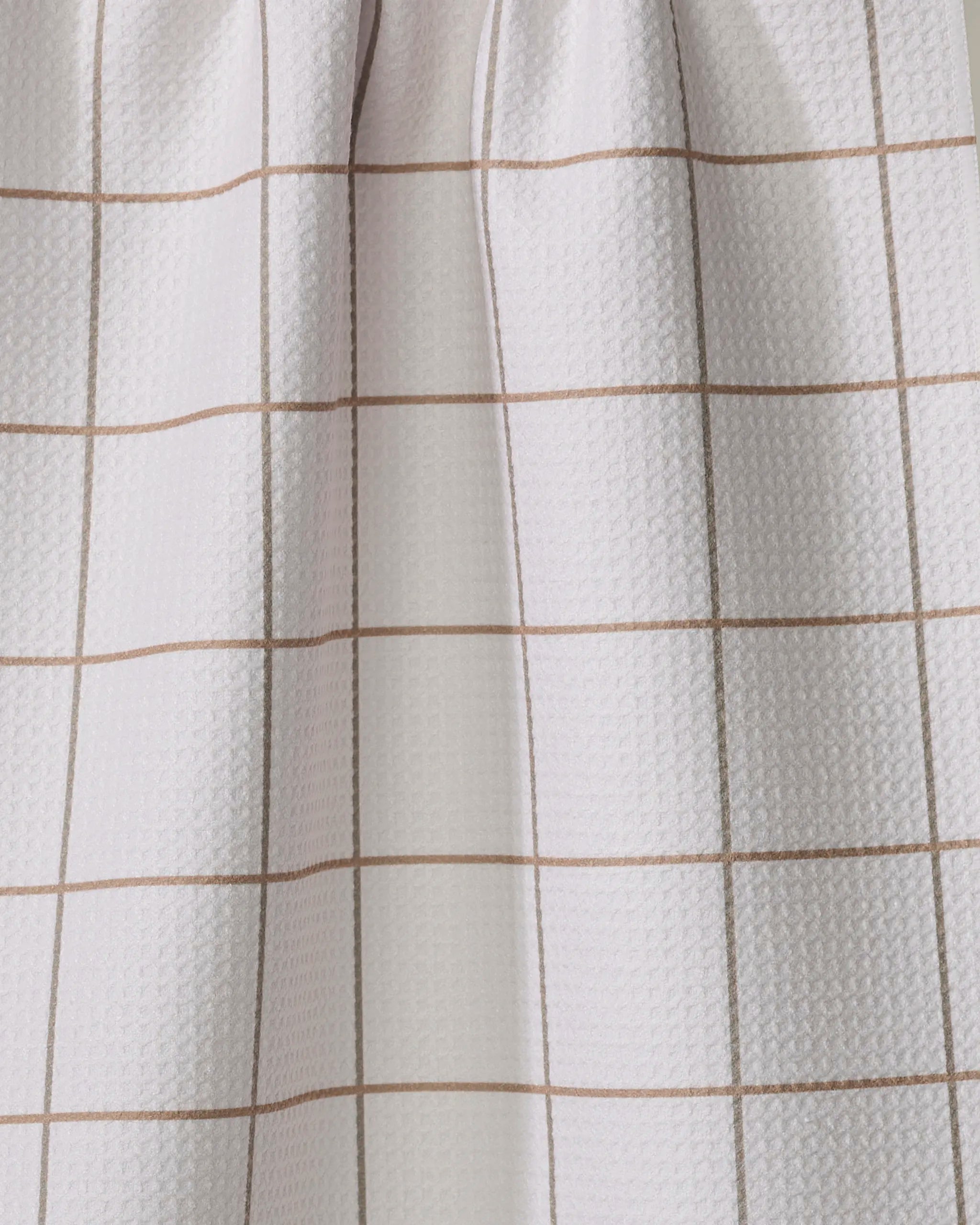 The Kelsi Fullmer Ivory Grid kitchen towel features a light-colored fabric with thin, evenly spaced brown lines in a grid pattern, draped to create soft folds and subtle shadows.