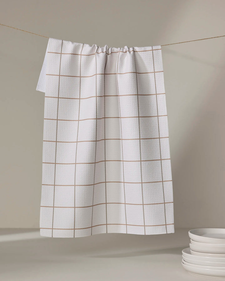 The Ivory Grid tea towel by Kelsi Fullmer, featuring a light brown grid on white, hangs on a line with stacked white plates in the corner against a neutral background.
