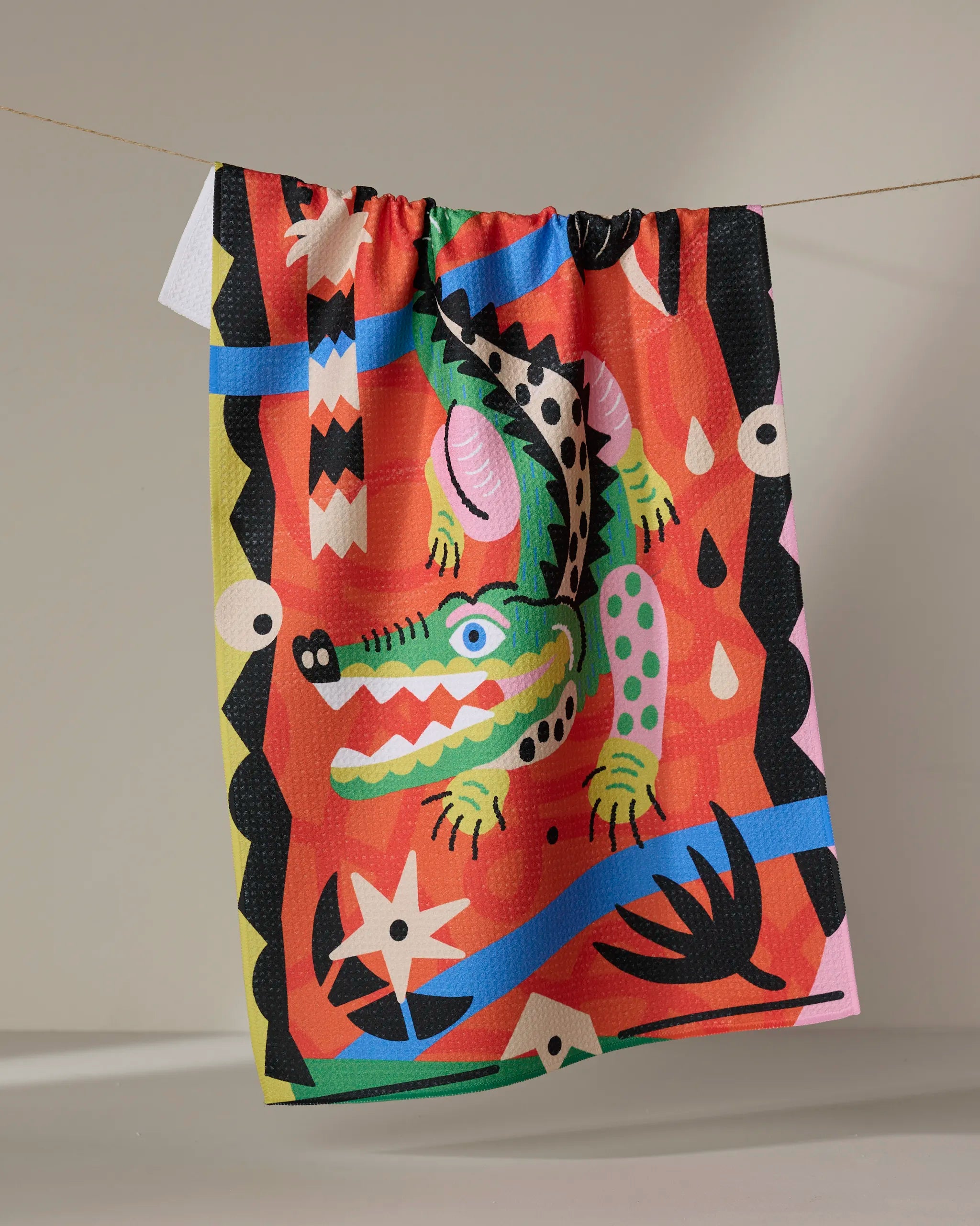 The Jacaré dish towel by Camila Leão features a playful crocodile illustration and abstract blue, pink, yellow, black, and white patterns. Colorful and absorbent, it hangs on a clothesline against a plain background.