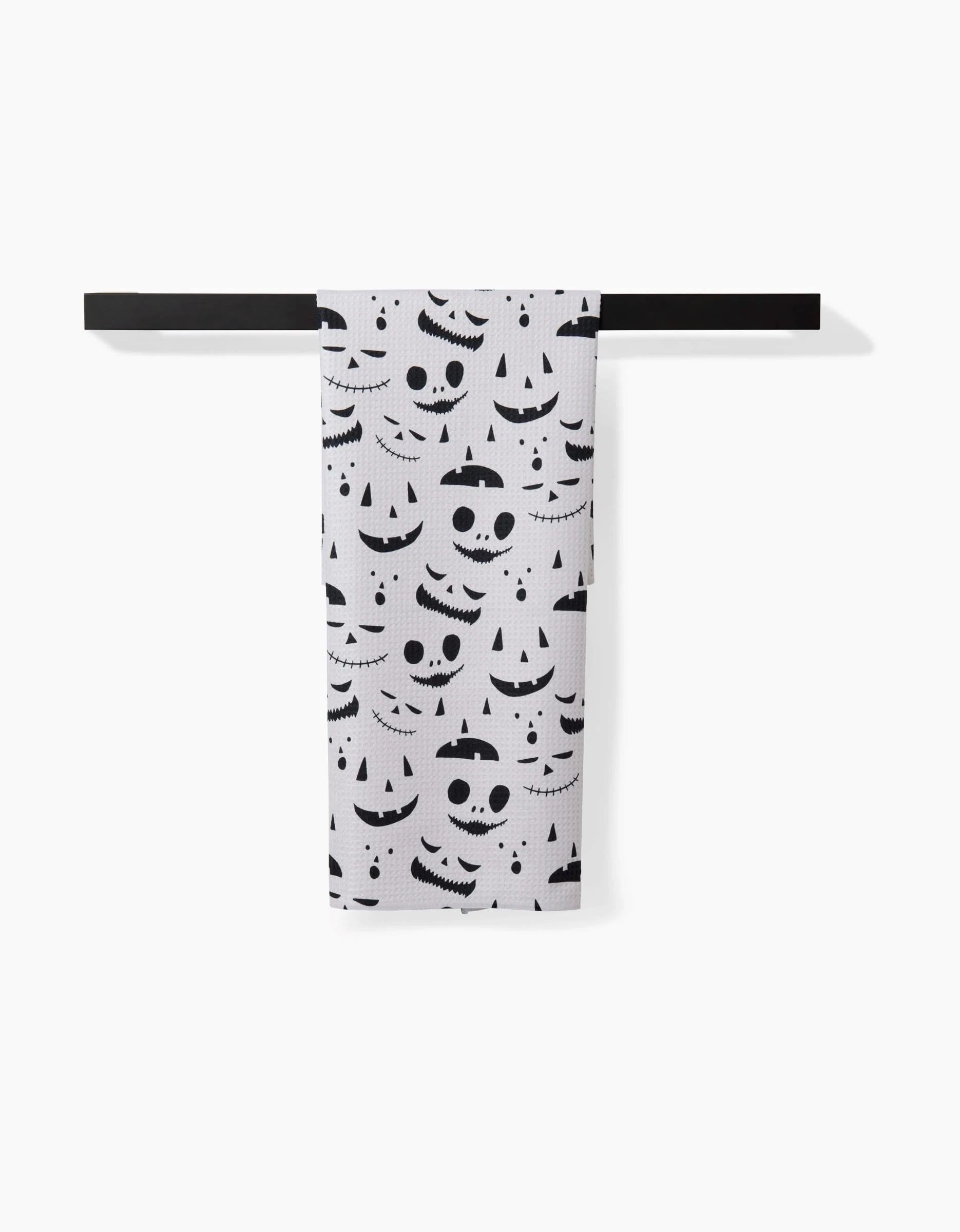 The Danelys Sidron Jack O Lantern Cream tea towel, featuring black Halloween jack-o’-lantern faces, hangs neatly on a modern black towel bar against a plain white background.