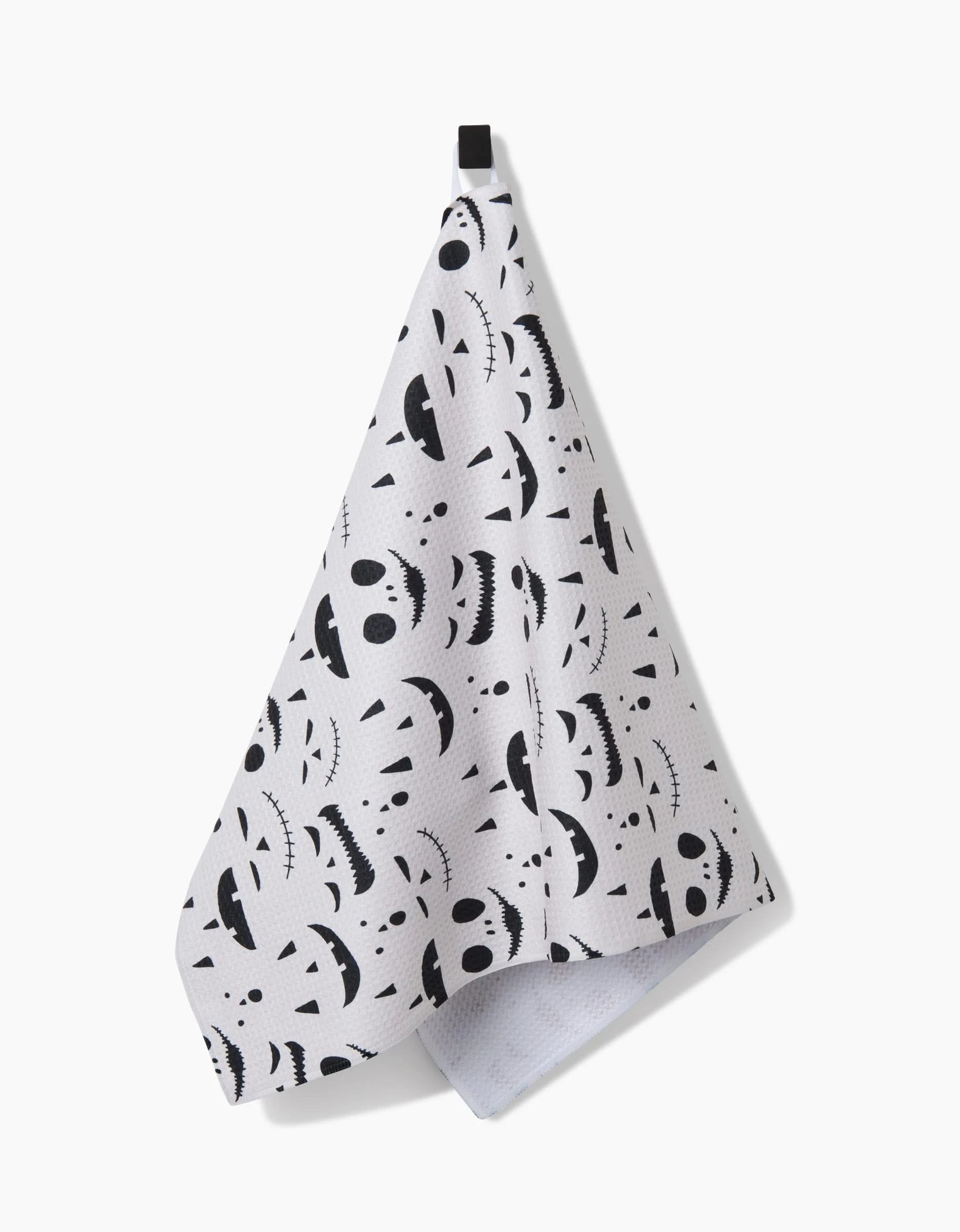 The Jack O Lantern Cream by Danelys Sidron is a white recycled kitchen towel with black Jack-o-lantern faces in various expressions, hung by a black loop against a plain white background.