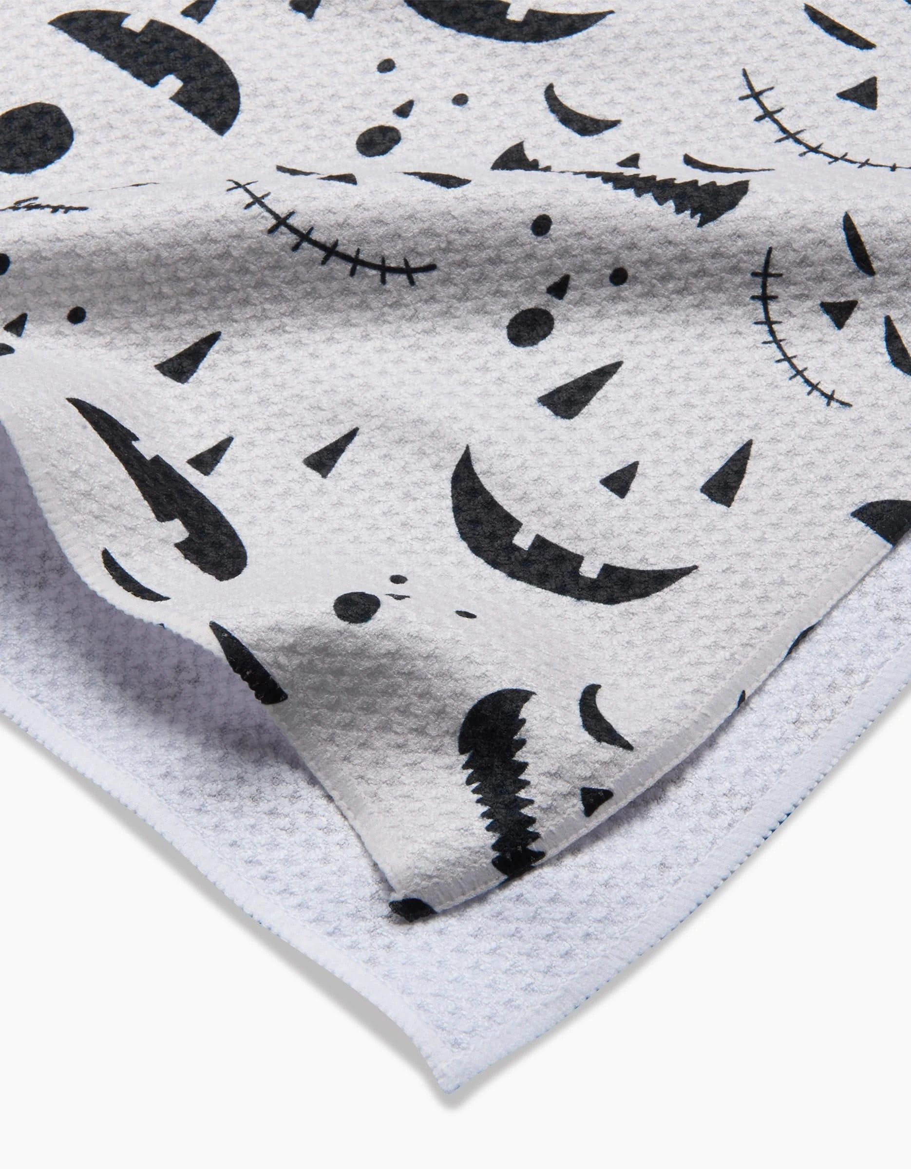 A close-up of the Danelys Sidron Jack O Lantern Cream tea towel, featuring a white textured fabric adorned with black Halloween-themed jack-o-lantern faces in smiling and spooky expressions.