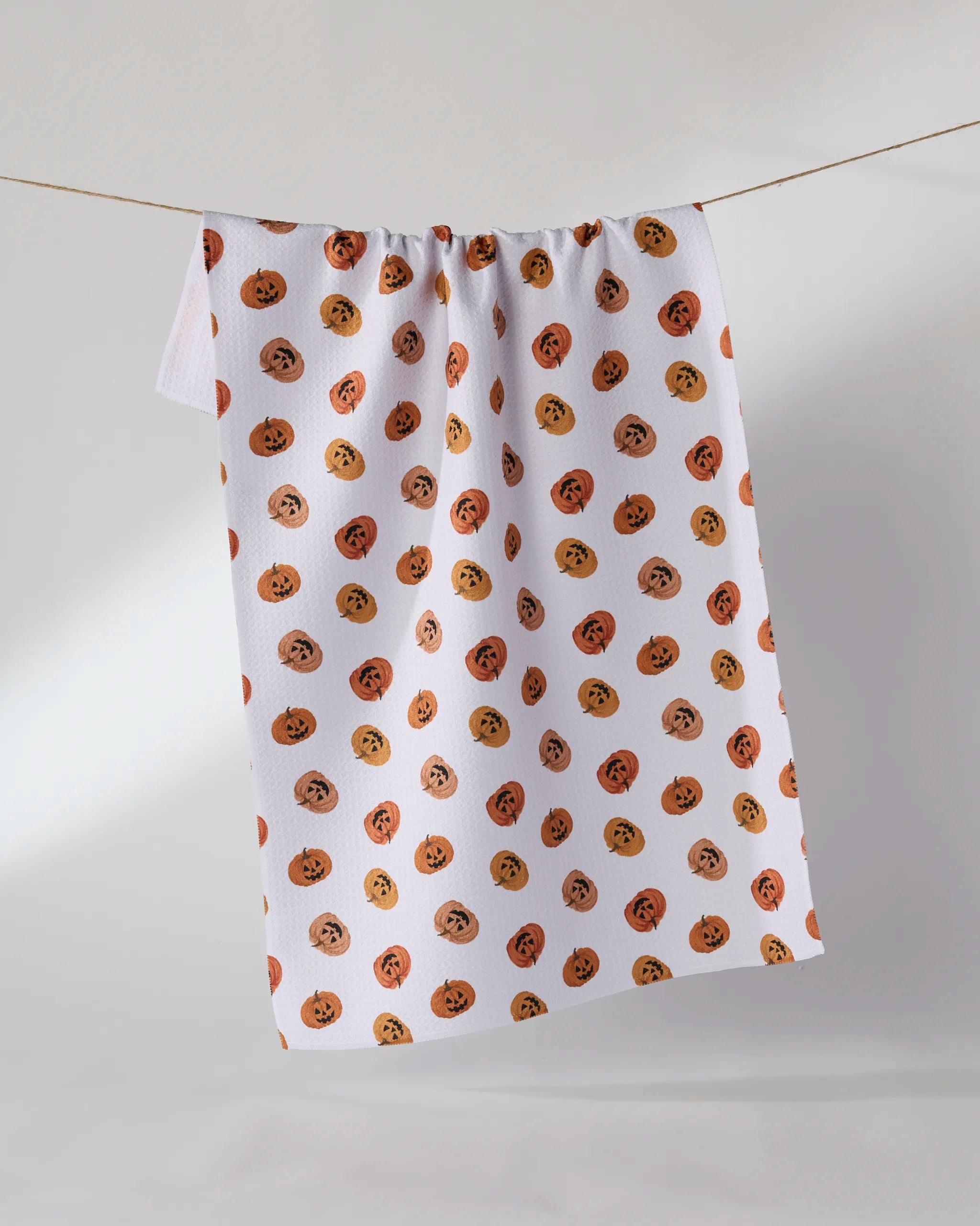 The Geometry Jacks Parade tea towel is ultra-absorbent and features a repeating pattern of smiling jack-o-lanterns in orange hues on a white background, shown hanging on a clothesline against a plain light backdrop.