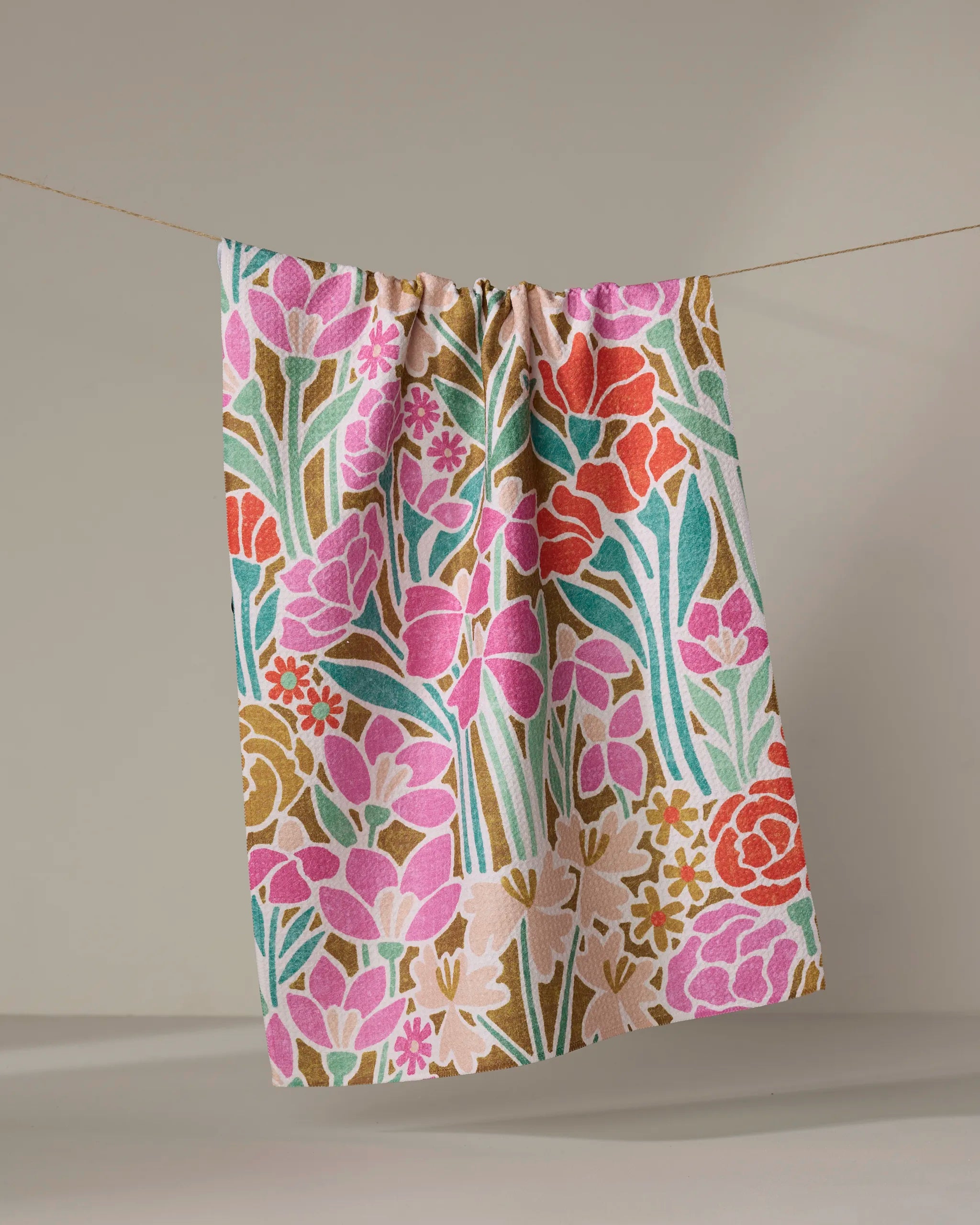 The Janae kitchen towel by Amy MacCready, featuring a vibrant floral pattern in pink, green, gold, and red, hangs on a clothesline against a neutral background.