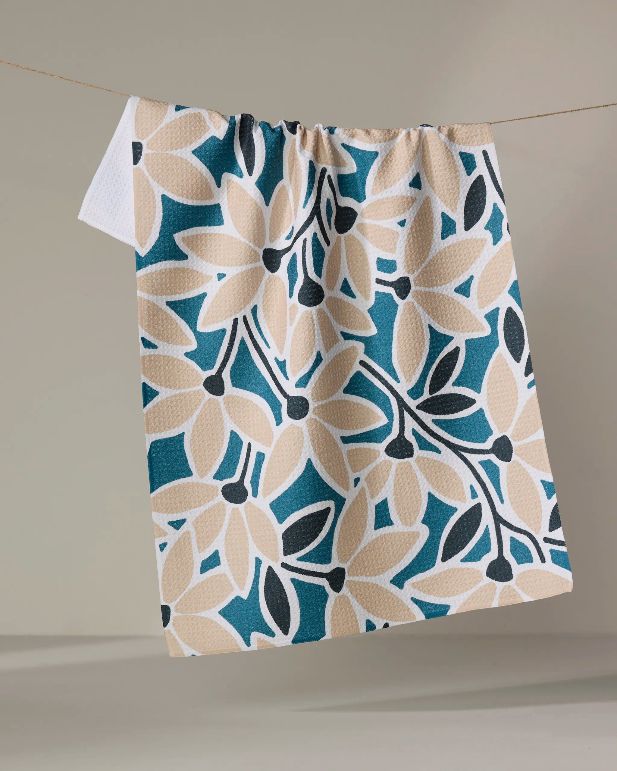 The Janine tea towel by Amy MacCready, featuring bold floral patterns in beige, dark blue, and teal, adds stylish function to your kitchen as it hangs beautifully against a neutral background.