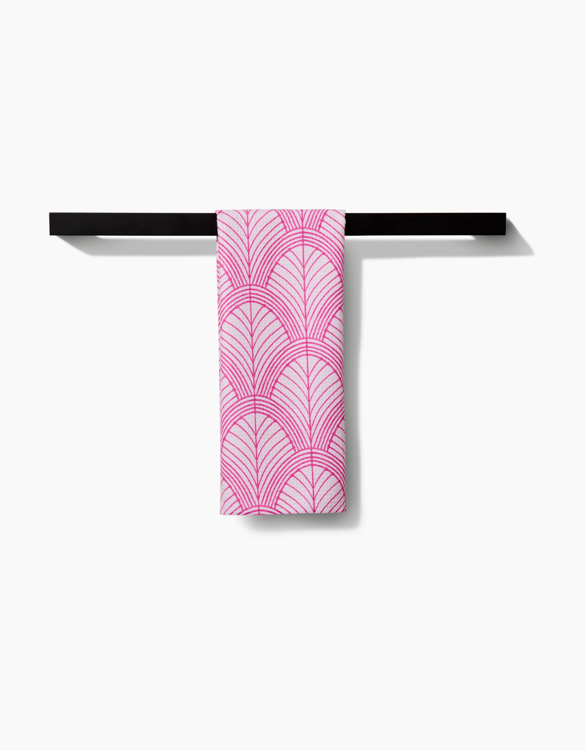 A pink and white Amy MacCready Jaskin tea towel featuring a repeating scallop shell pattern hangs neatly over a simple black towel bar against a plain white background.