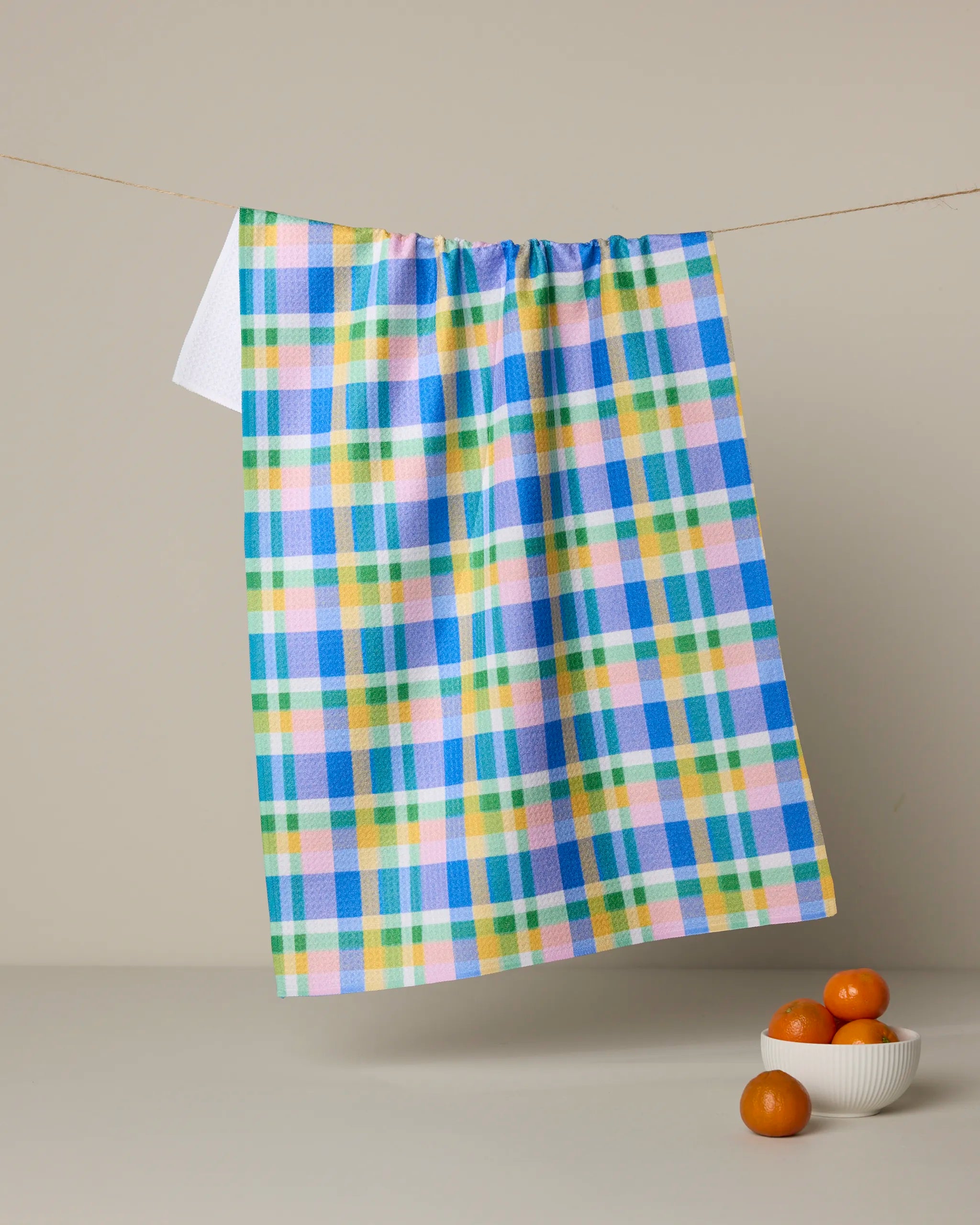 The Geometry Jelly Bean tea towel, featuring blue, green, yellow, and pink stripes, hangs on a line above a white bowl of small oranges on a light surface with a neutral background.
