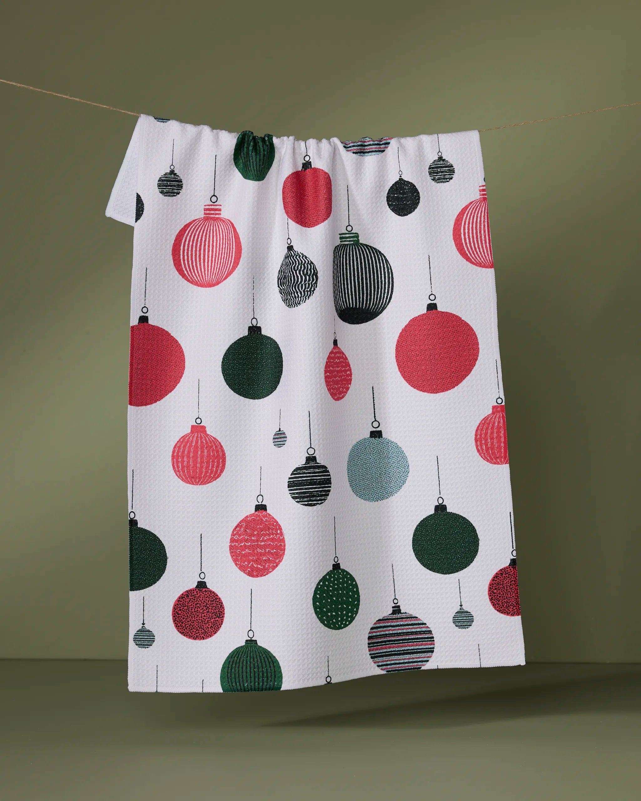 The Geometry "Jingles" tea towel, white and ultra-absorbent with red, green, and black Christmas ornament designs, hangs on a line against an olive green background.