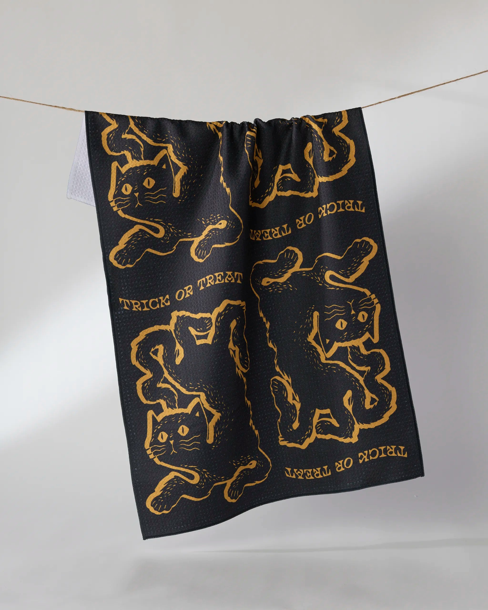 The Geometry Jinx tea towel, made from ultra-absorbent recycled materials, displays yellow cat illustrations and repeating Trick or Treat text, hanging on a clothesline against a plain light backdrop.