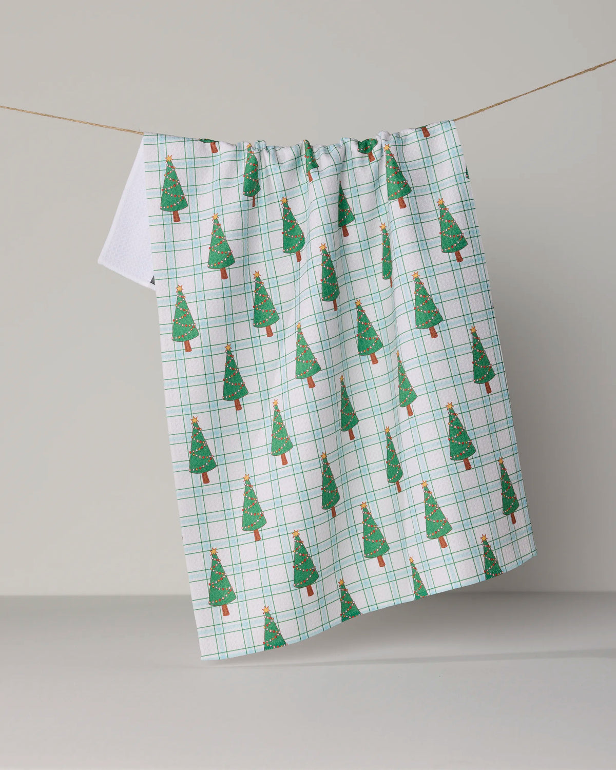 The Geometry Jolly Plaid kitchen tea towel features a green and red plaid pattern adorned with Christmas trees, hanging on a string against a plain backdrop. Made from recycled materials, this ultra-absorbent and eco-friendly towel brings festive charm and practical style to your home.