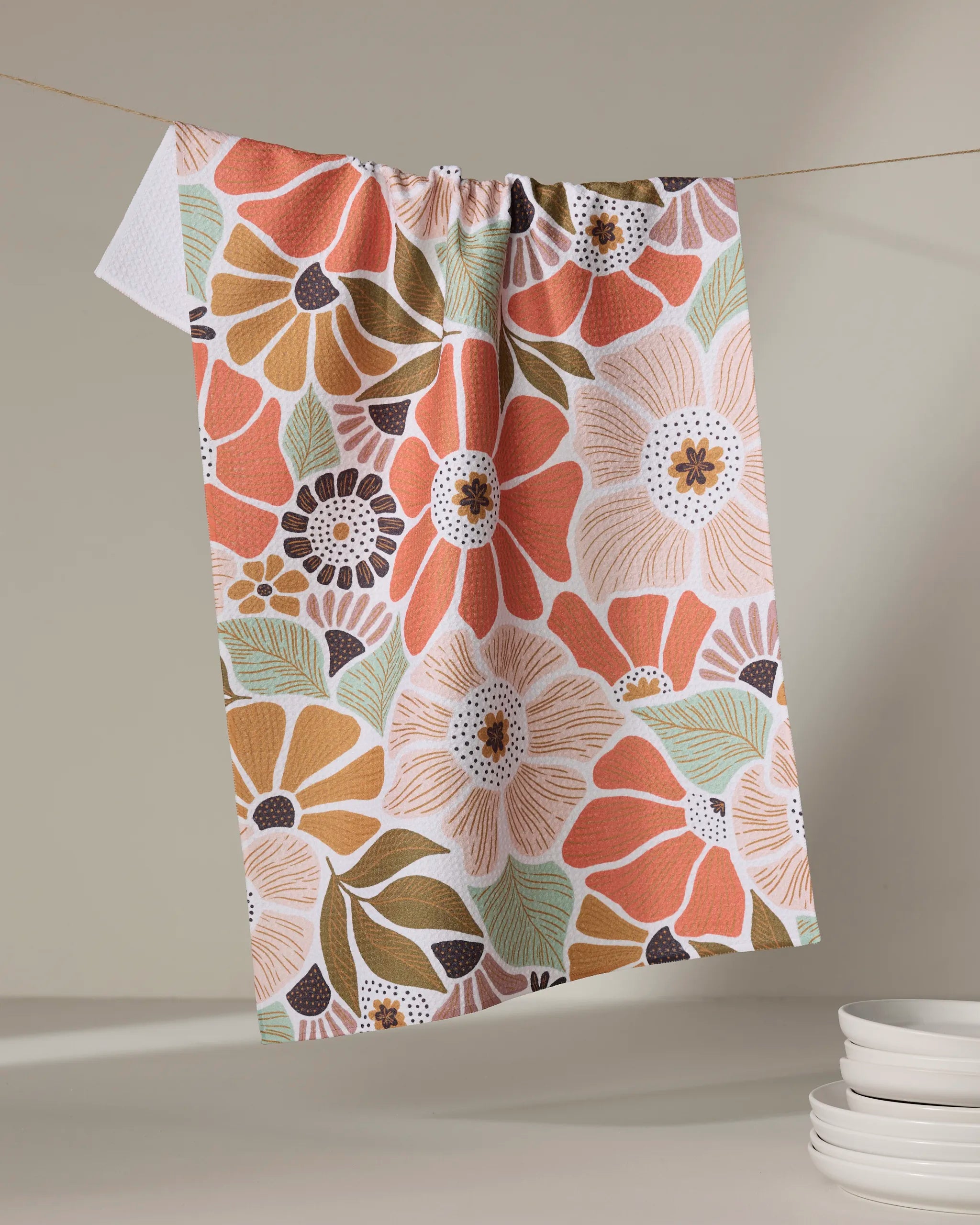 The Jorah tea towel by Amy MacCready, featuring a bold floral pattern in orange, pink, yellow, and green, hangs from a string while white plates are stacked nearby against a neutral background.