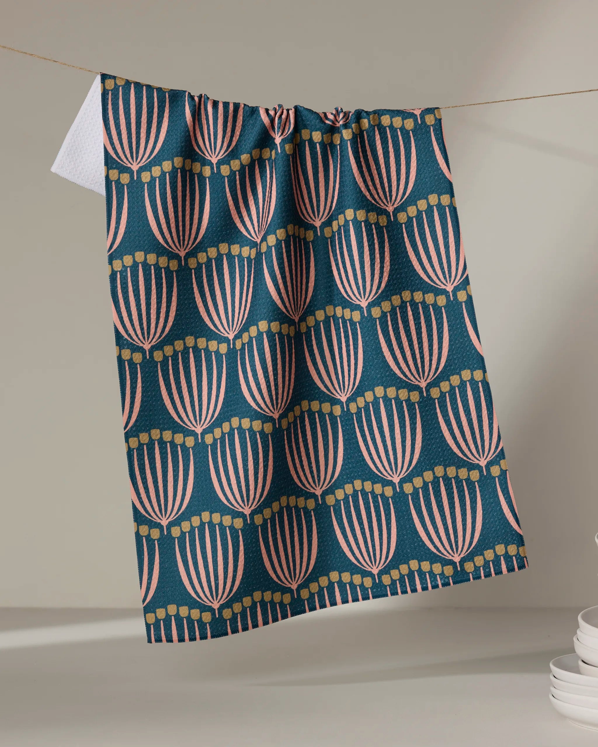 The "Josie" towel by Amy MacCready, featuring a dark teal eco-friendly fabric with pink and yellow floral patterns, hangs from a string indoors. White plates are neatly stacked in the bottom right corner.