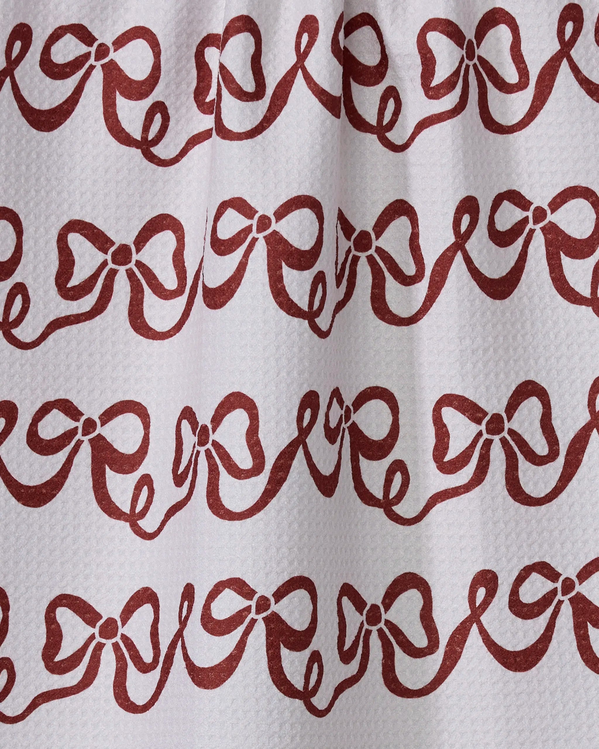 The Geometry Joyful Bows tea towel is white and textured, made from absorbent recycled materials, featuring a repeating pattern of red bows and wavy horizontal lines.