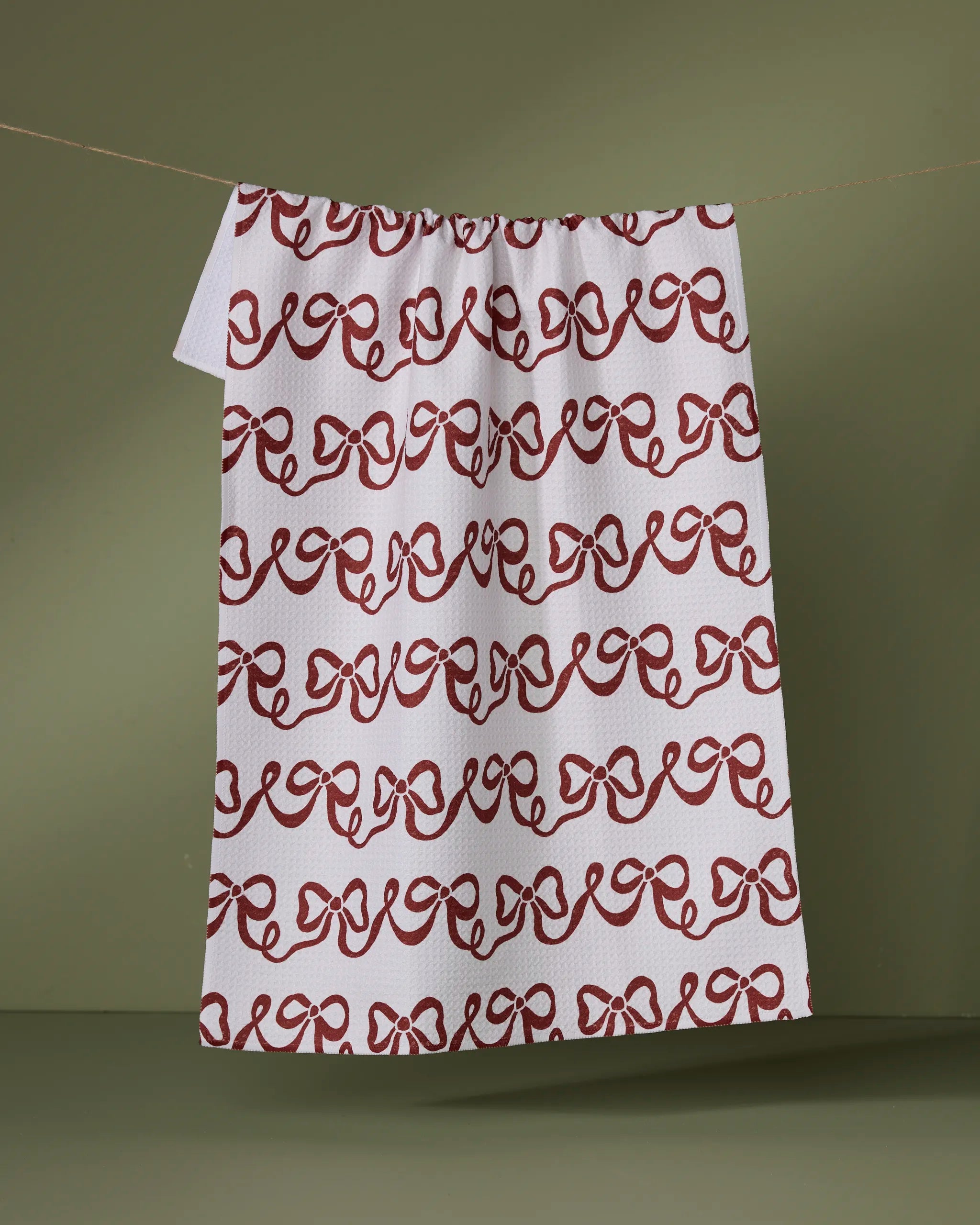 The Geometry Joyful Bows tea towel, featuring a repeating red bow pattern on white, hangs against an olive green background. Made from absorbent recycled materials, it’s both stylish and eco-friendly.