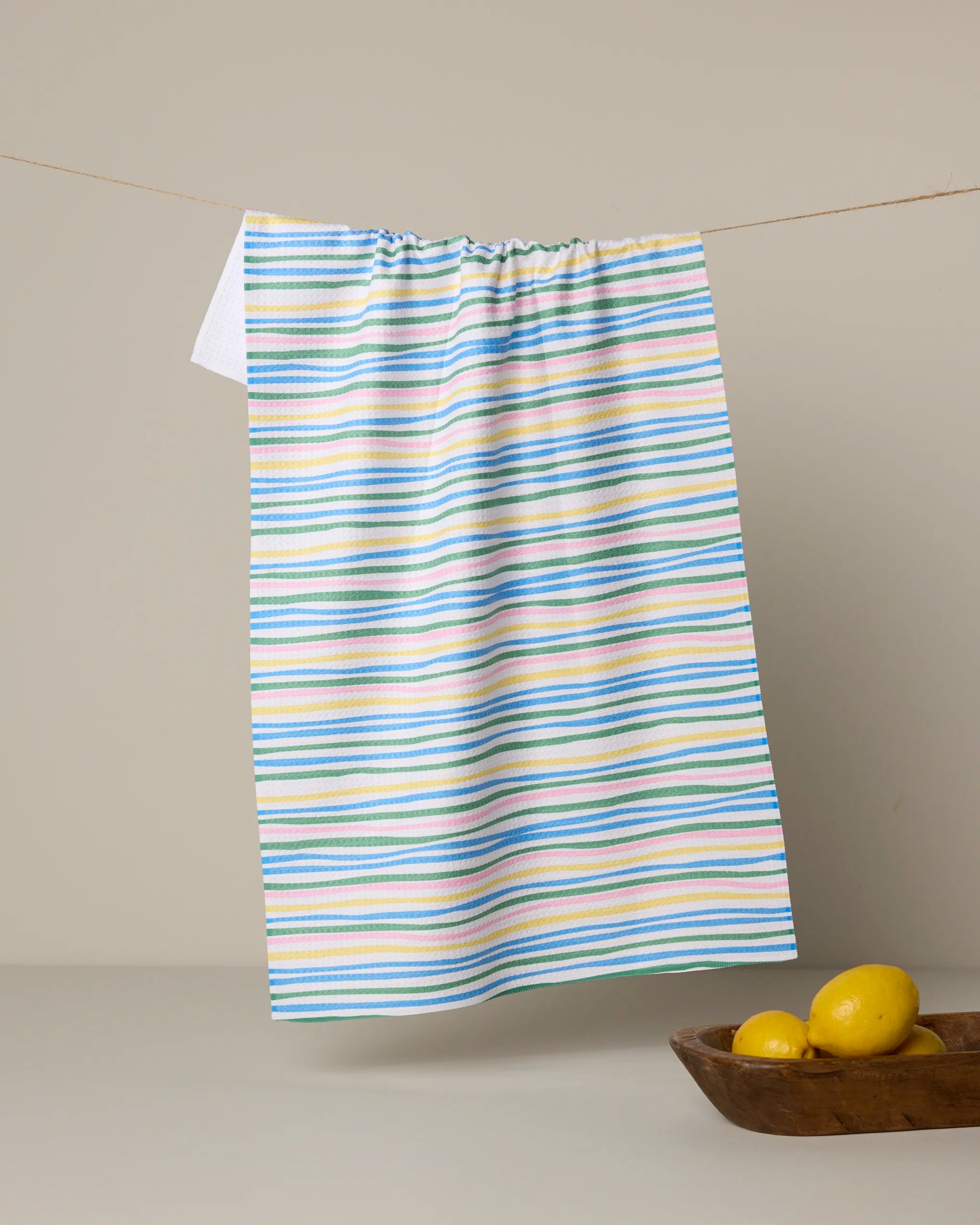 The Jubilation kitchen towel by Geometry, featuring pastel blue, yellow, pink, and green stripes, hangs on a string with a wooden bowl of lemons below, set against a neutral background.