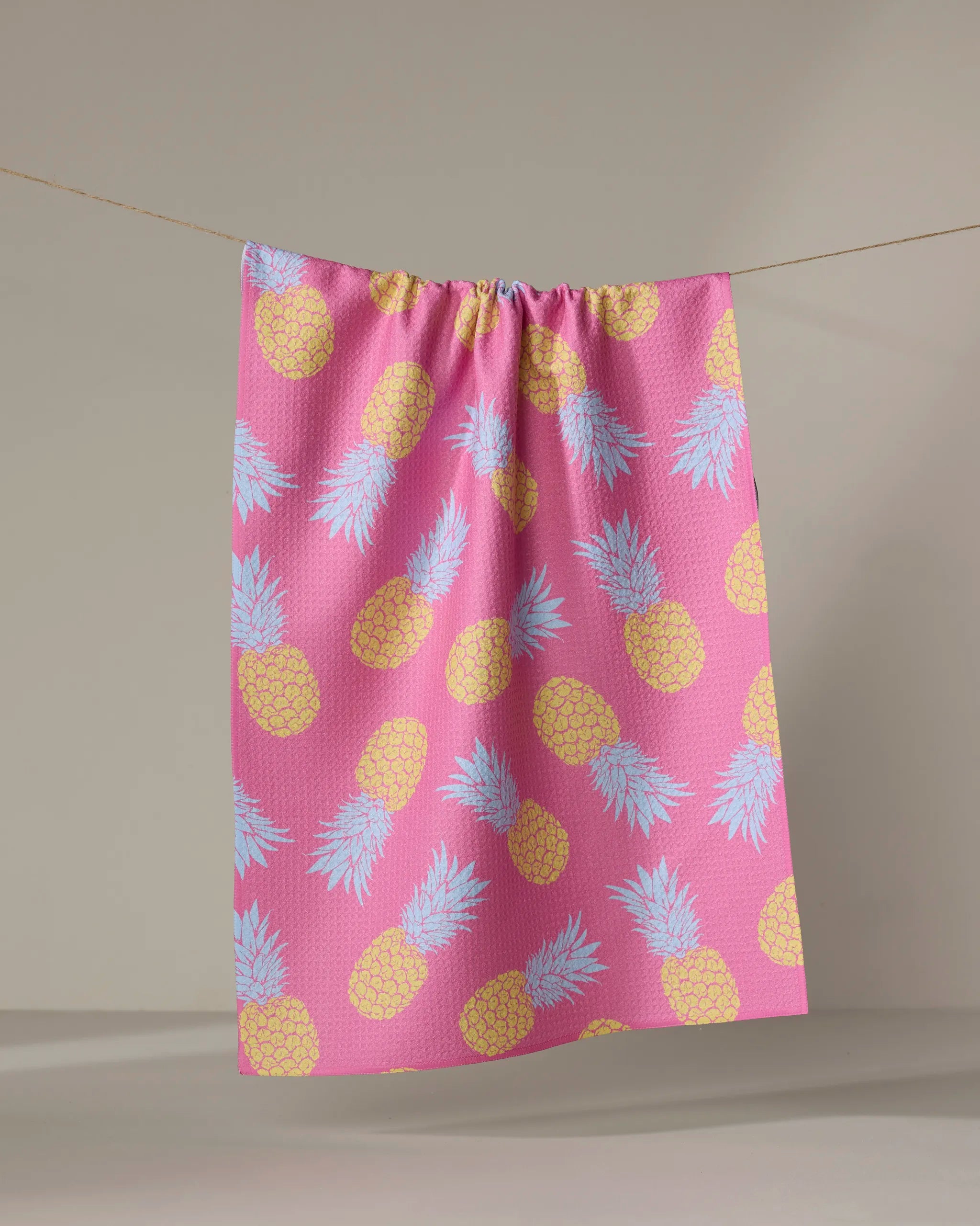 The "Juicy Pineapples" kitchen towel by Ashley Celena features recycled pink fabric with yellow pineapples and light blue leaves, hanging on a line against a simple, light background.