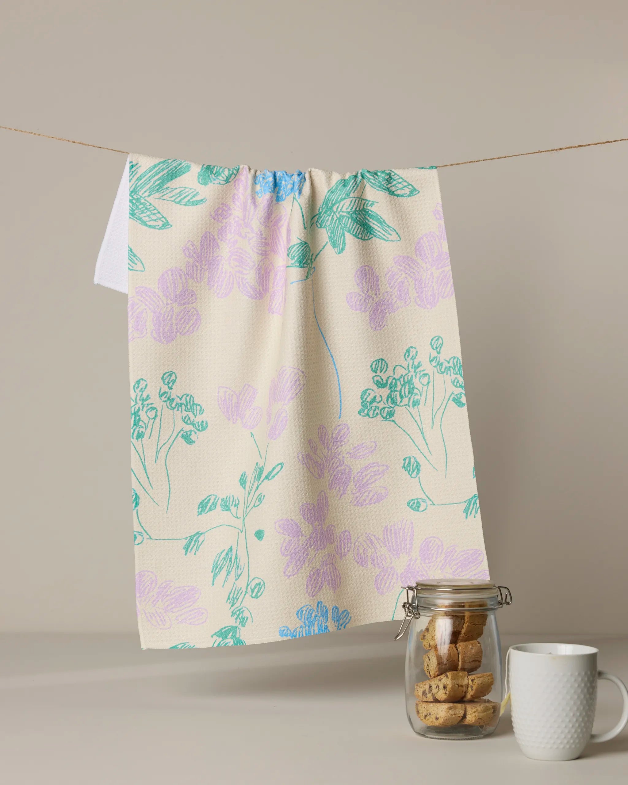 The Julieta tea towel by Geometry, featuring pastel floral patterns and quick-drying fabric, hangs on a line. Below, a glass jar of cookies and a white textured mug rest on a light surface with a neutral backdrop.
