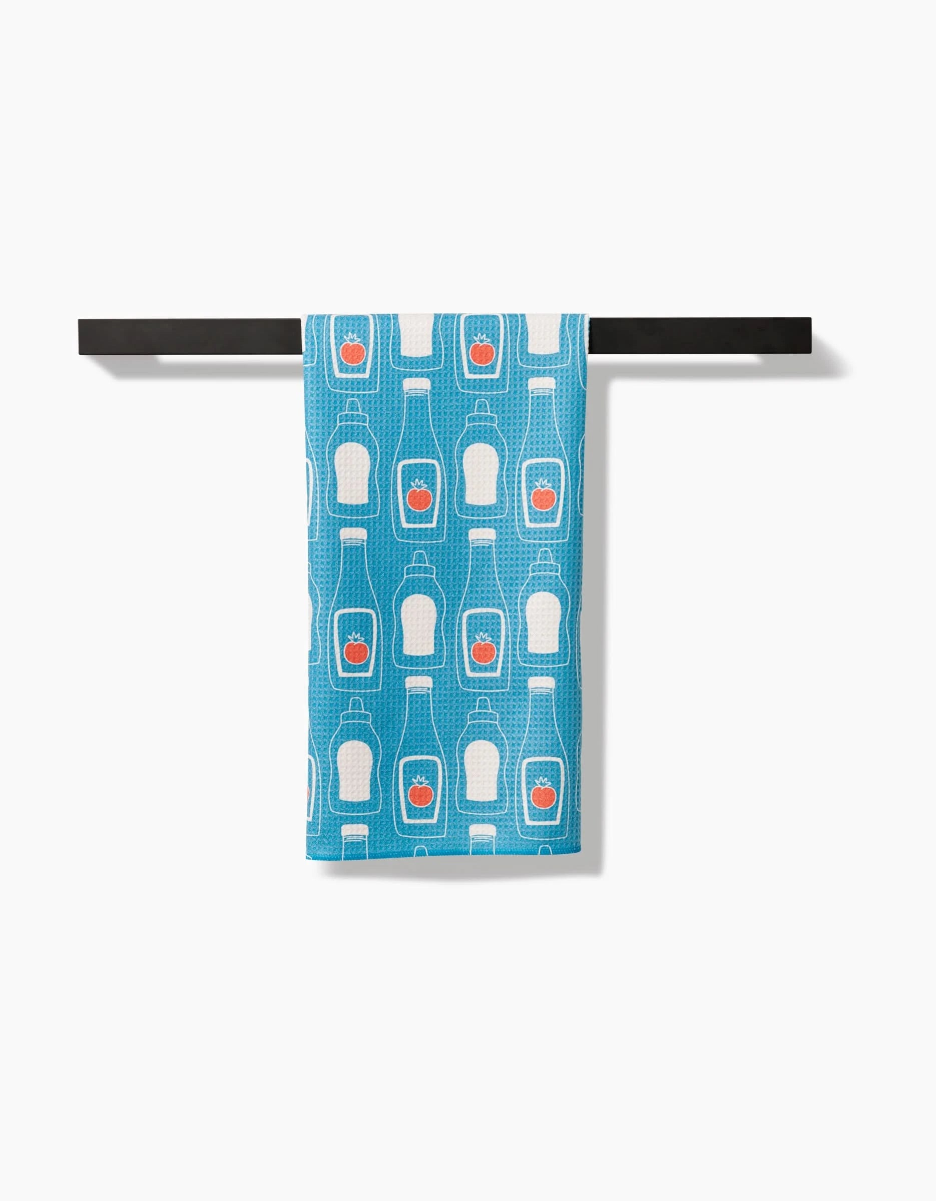 The July Squeeze kitchen towel by Sara Mari features ultra-absorbent blue Geoweave fabric with a white ketchup bottle pattern and red labels, shown draped over a black rod on a white background.