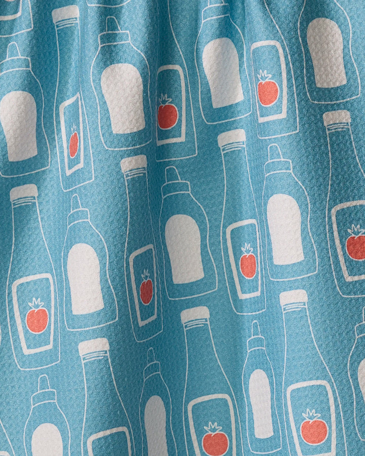 July Squeeze" by Sara Mari features blue fabric with playful white-outlined condiment bottles, some with red tomatoes—a charming, eco-friendly kitchen towel that blends style and practicality.