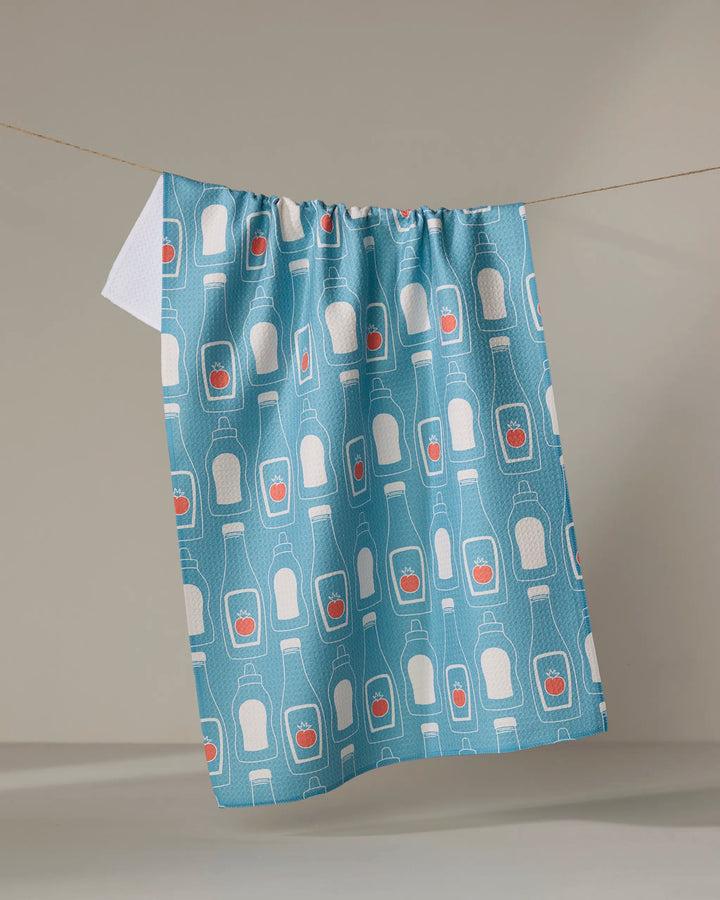 The July Squeeze by Sara Mari is an eco-friendly blue kitchen towel with white bottles and orange hearts, hanging on a clothesline against a neutral background.