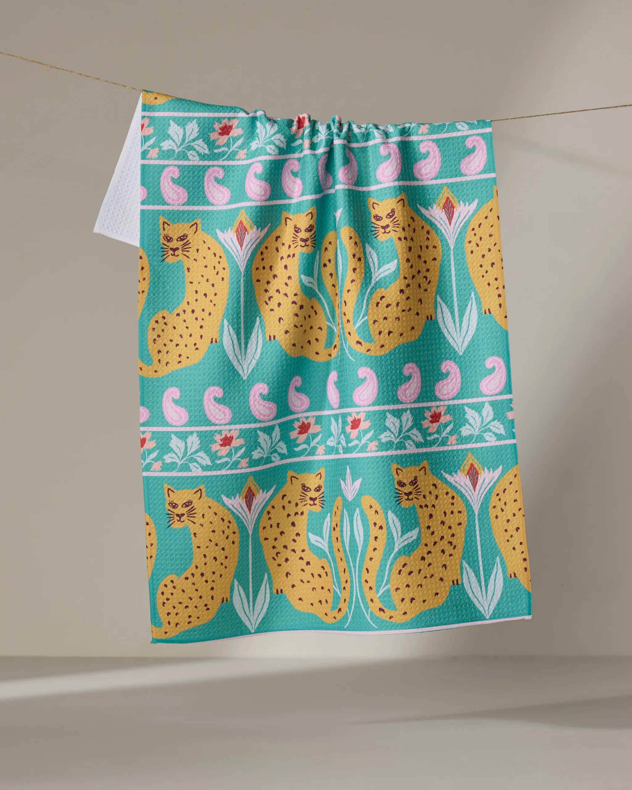 The Jungle Blooms tea towel by Jasmine Blooms Designs hangs on a line, displaying yellow cheetahs, pink and green flowers, and paisleys on turquoise—crafted from recycled materials.