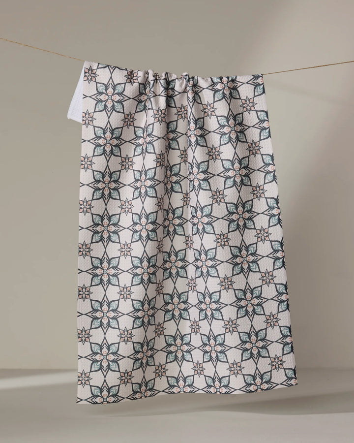 The "Jungle Night" kitchen towel by Amita Nair, featuring a blue, green, and brown geometric floral print, hangs gathered on a string against a neutral beige background—an eco-friendly way to add charm to your space.