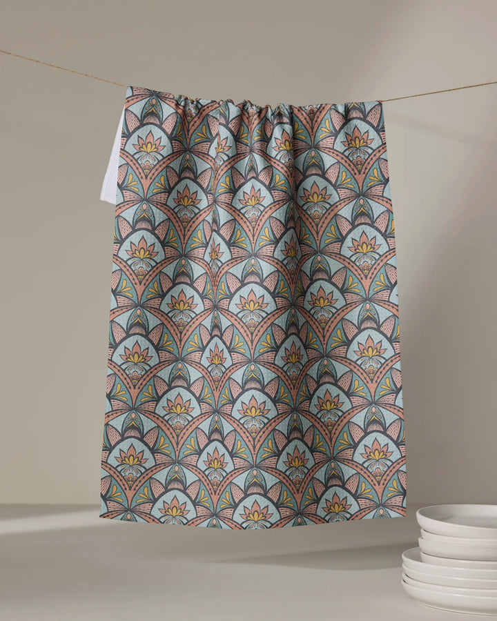 Amita Nair's "Jungle Sunrise" tea towel, made from recycled materials and ultra-absorbent, features vibrant geometric and floral patterns. It hangs by white plates stacked against a softly lit neutral background.