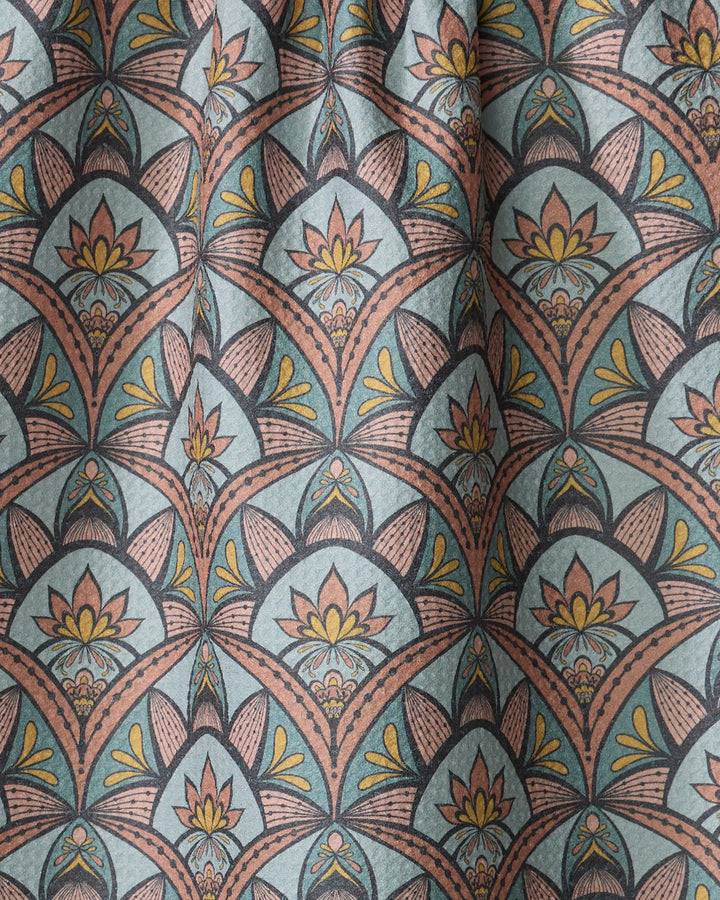A close-up of the "Jungle Sunrise" tea towel by Amita Nair, made from recycled materials. It features a repeating art deco floral pattern in yellow, peach, and black on a blue-green background with gentle folds and slight texture.