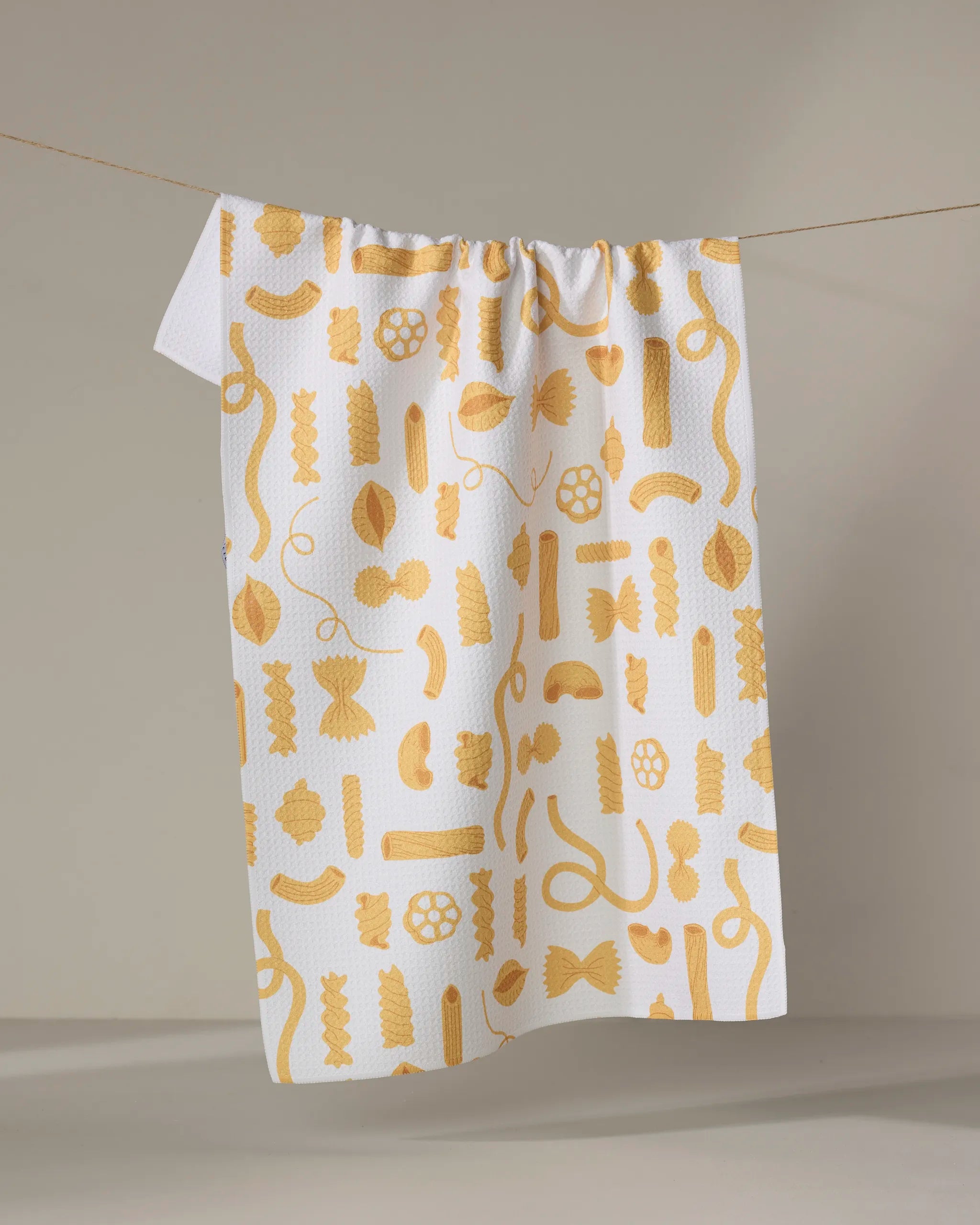 The Just Add Sauce tea towel by Vaishali Design Studio, ultra-absorbent and white with yellow pasta shapes, hangs on a clothesline against a plain light background.