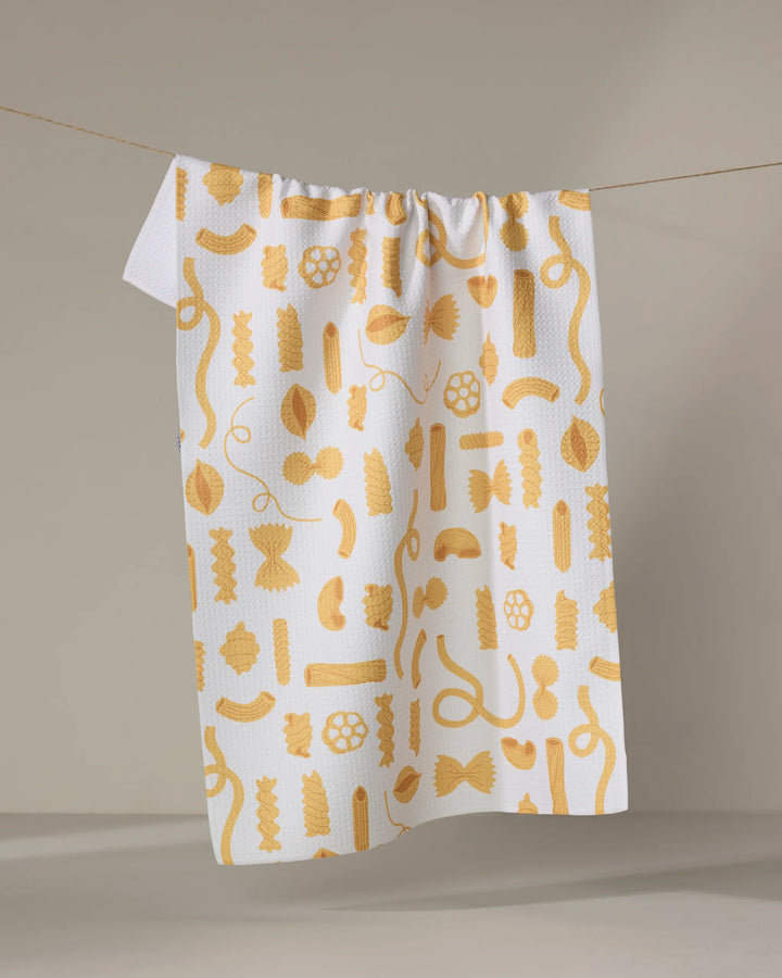 The Just Add Sauce tea towel by Vaishali Design Studio, ultra-absorbent and white with yellow pasta shapes, hangs on a clothesline against a plain light background.