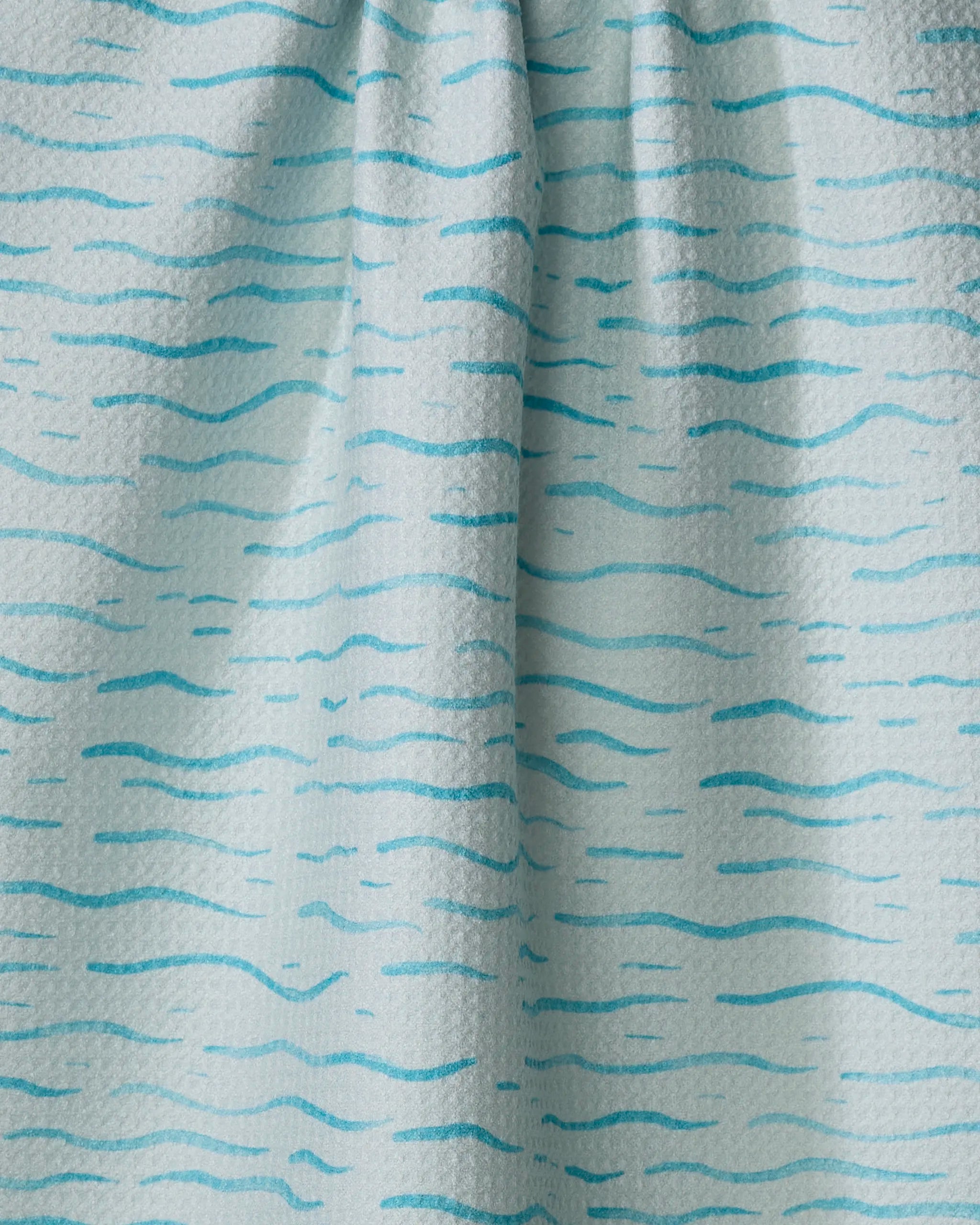 Close-up of the Just Keep Swimming tea towel by Katie O'Shea, featuring light blue fabric with irregular turquoise lines like water waves. The center is softly gathered, creating gentle folds and shadows.