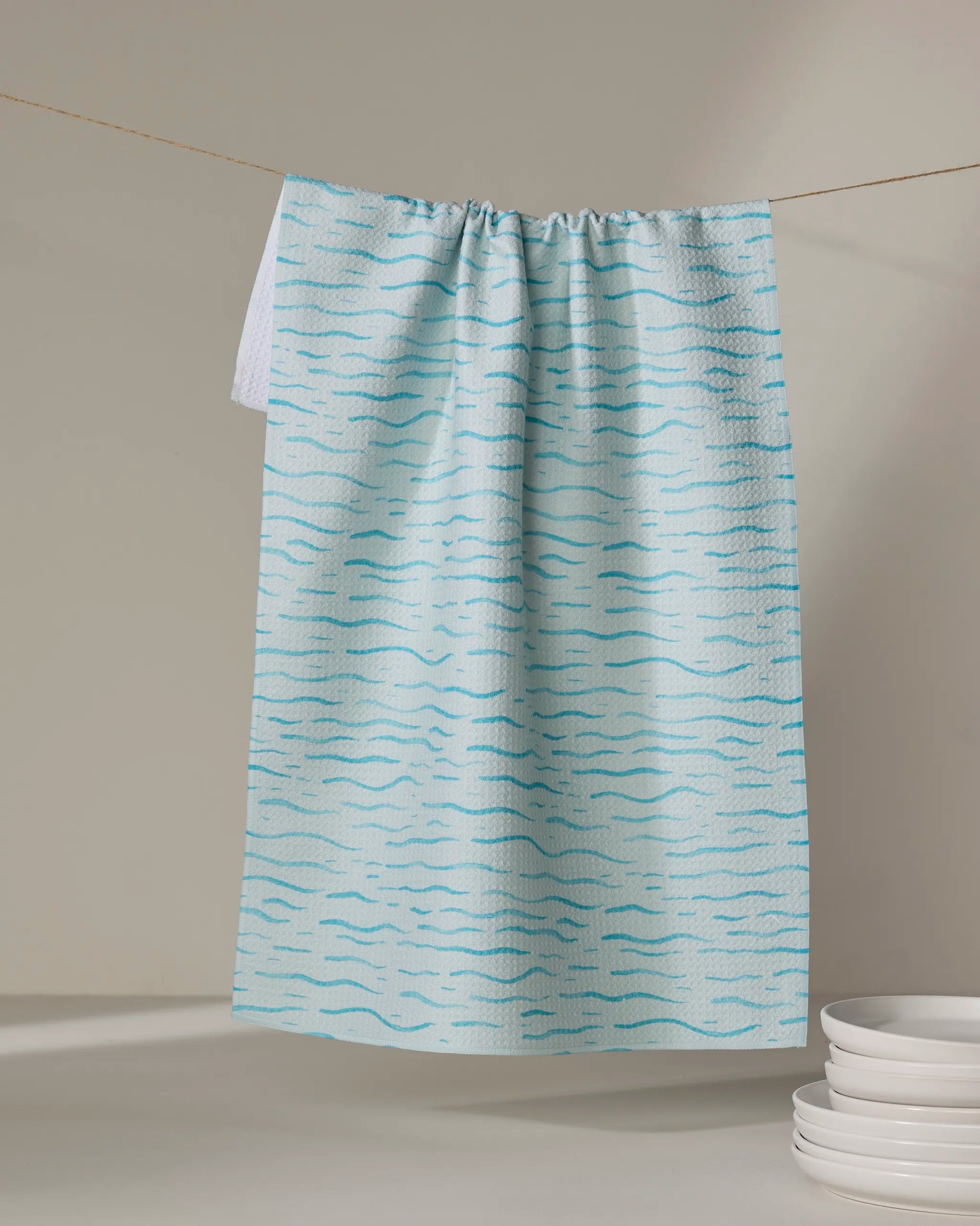 The Katie O'Shea "Just Keep Swimming" light blue quick-drying tea towel with a wavy blue line pattern hangs on a string near a stack of white plates, set against a neutral background.