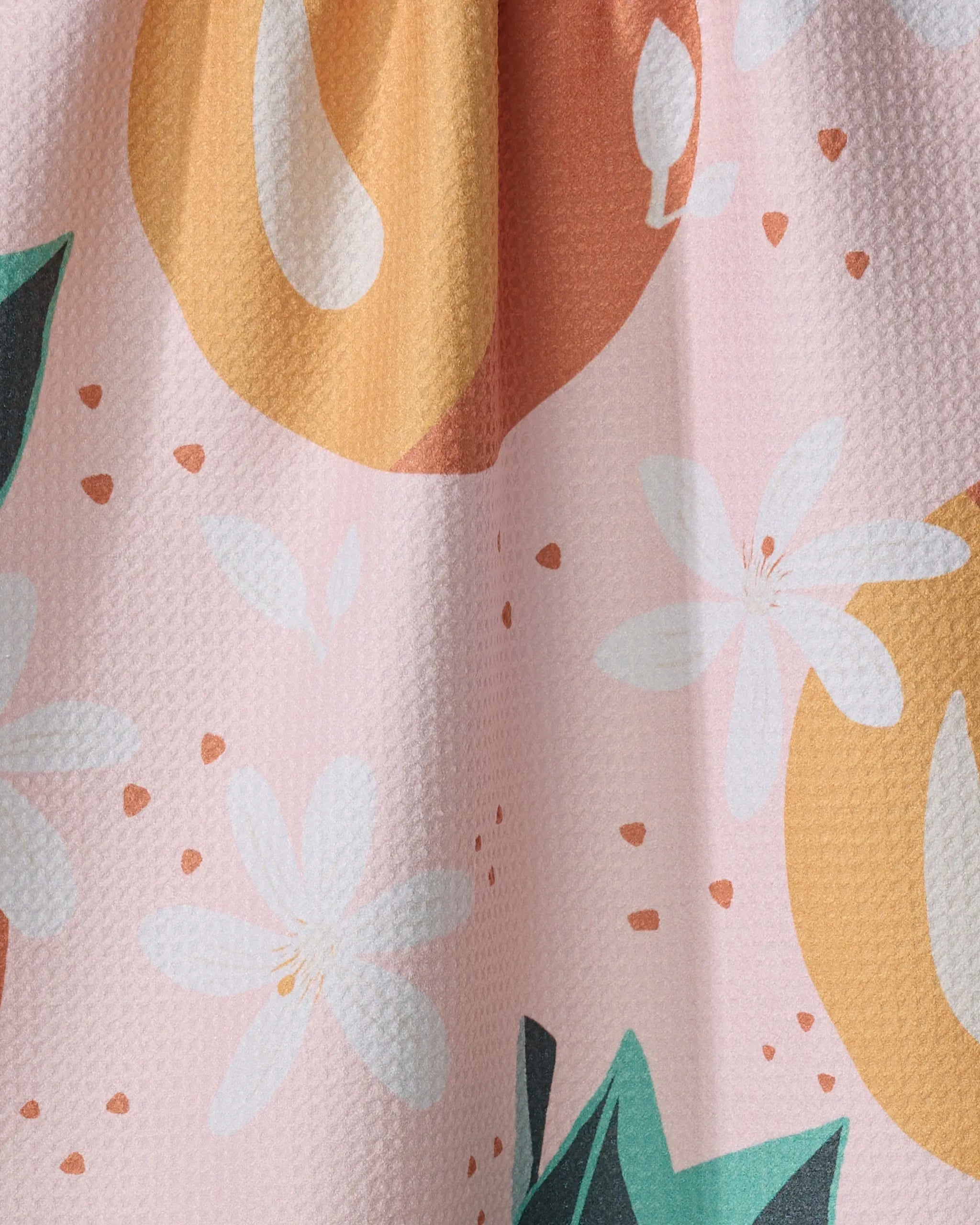 A close-up of the Just Peachy recycled kitchen towel by Julianne Haness, featuring white flowers, green leaves, orange fruit shapes, and small orange dots on a light pink textured background.