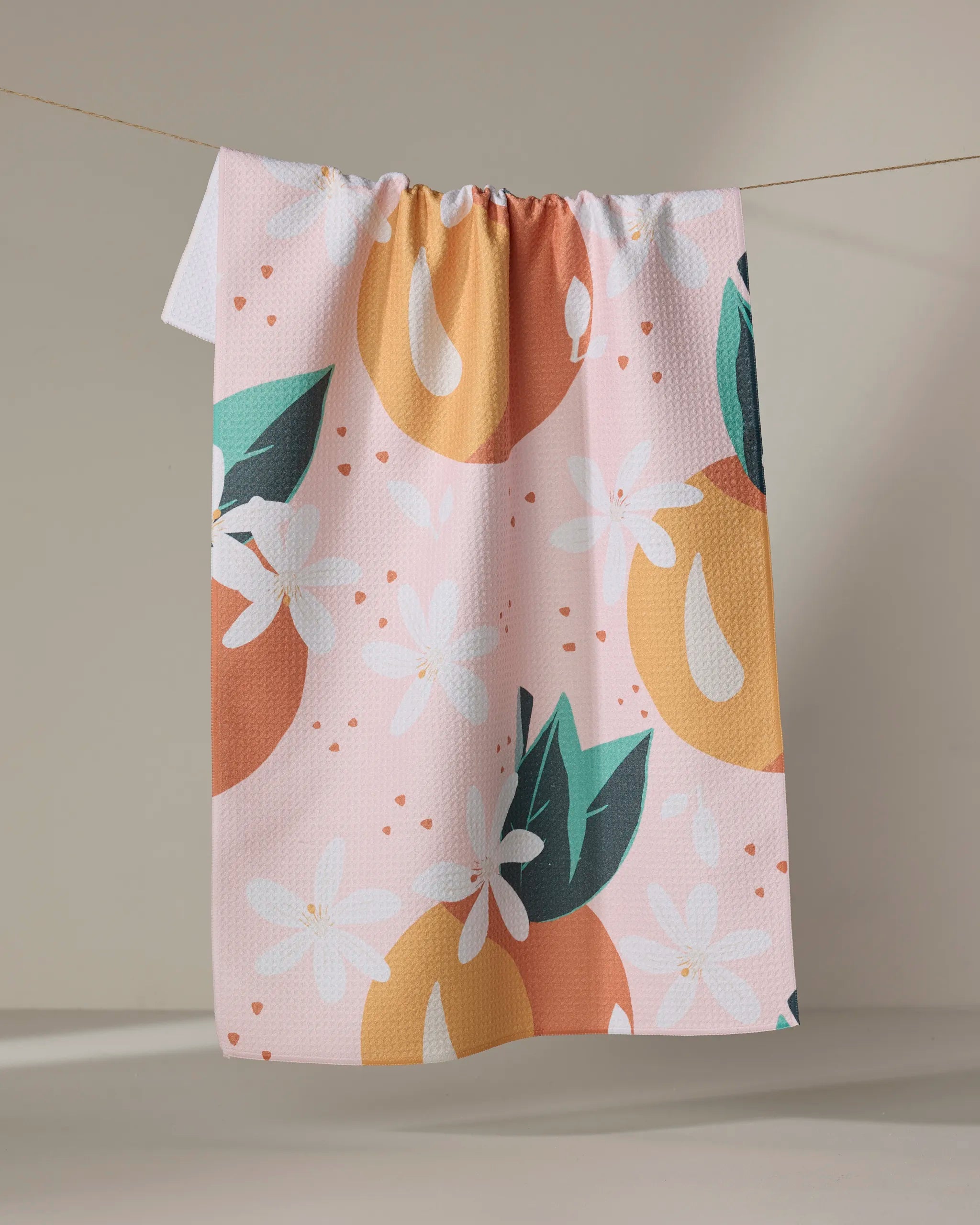 The "Just Peachy" tea towel by Julianne Haness, featuring large orange fruits, green leaves, and white flowers on a pink background, hangs on a clothesline against a neutral backdrop.