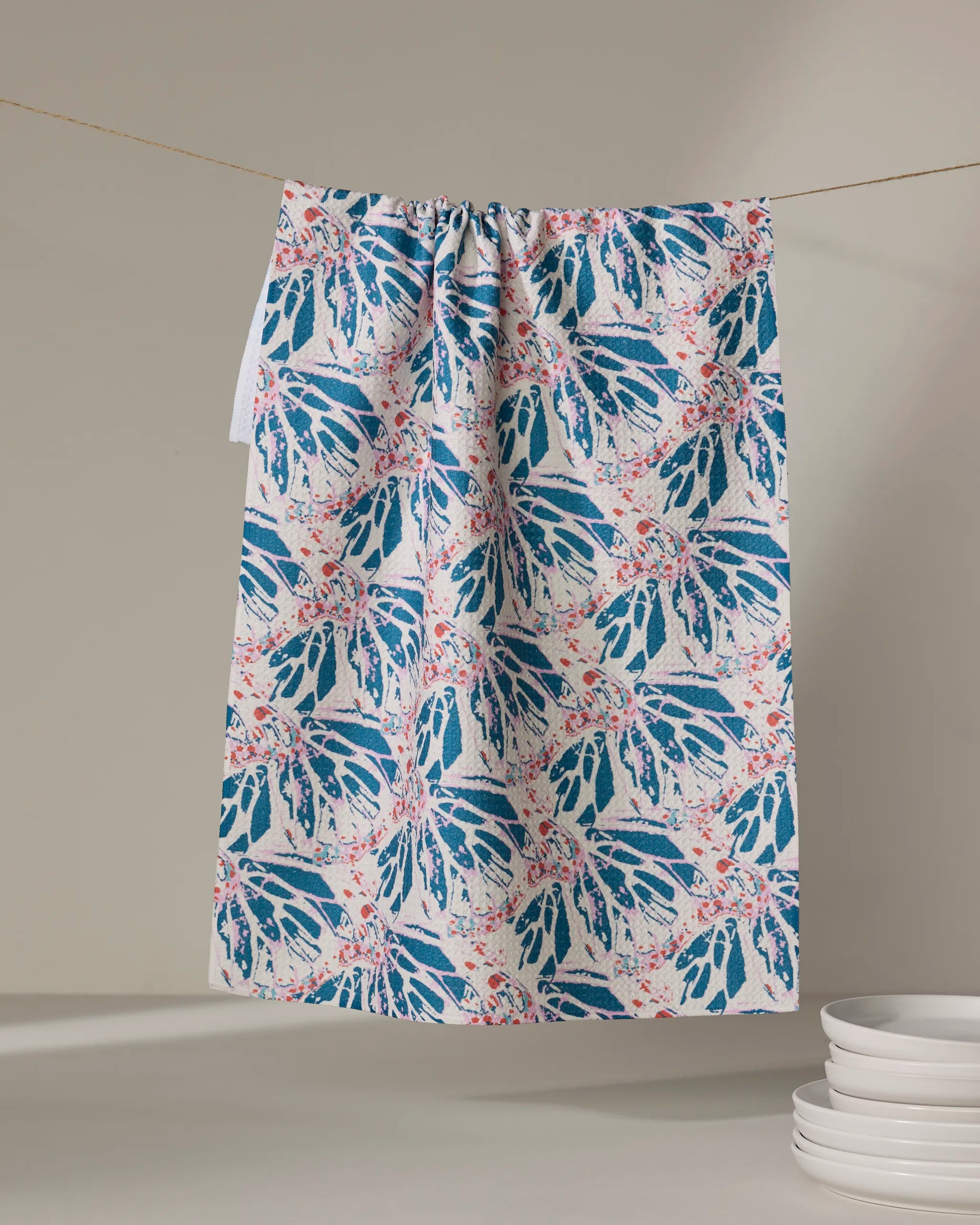 Rebecca Woolbright's "Kaleidoscope Dream" kitchen towel, featuring a blue and coral abstract leaf pattern, hangs on a clothesline with a stack of white plates nearby.