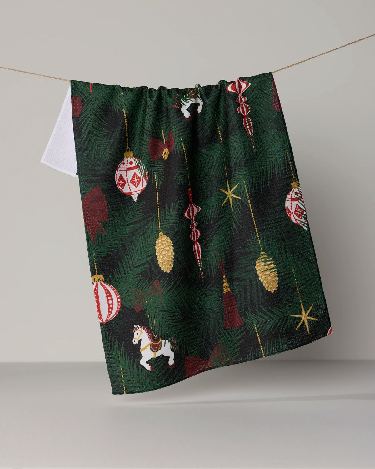 The Keepsake tea towel by Geometry, crafted from recycled materials, features green pine branches, gold pinecones, red and gold ornaments, and a white carousel horse in a festive design. Ultra-absorbent and environmentally friendly, it brings both holiday cheer and sustainability to your kitchen as it hangs on a clothesline against a plain background.